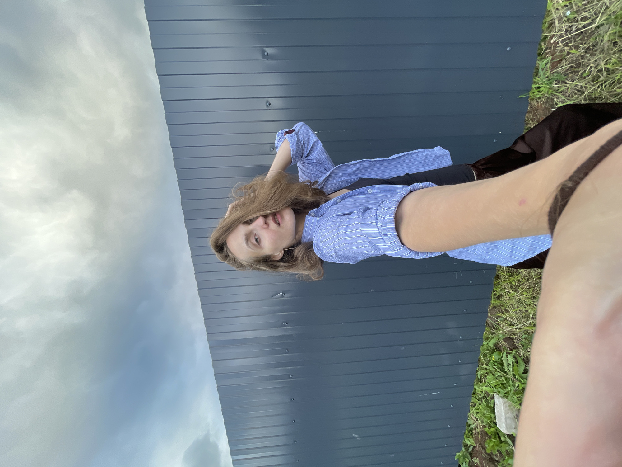 A young woman is lying on the grass in front of a dark blue metal wall, with her head resting on her hand. She is wearing a light blue striped button-up shirt, which is unbuttoned, revealing a dark garment underneath. Her hair is a light brown color and falls over her face and shoulders. The sky above is cloudy and appears to be overcast. To the right of the woman, there is a patch of green grass with some weeds and a small white object, possibly a piece of rock. The scene suggests an outdoor setting, possibly in a rural or suburban area, given the grass and simple wall. The image is taken from a low angle, looking up towards the sky. The lighting is soft, indicating it might be late afternoon or an overcast day. There is no visible text in the image.