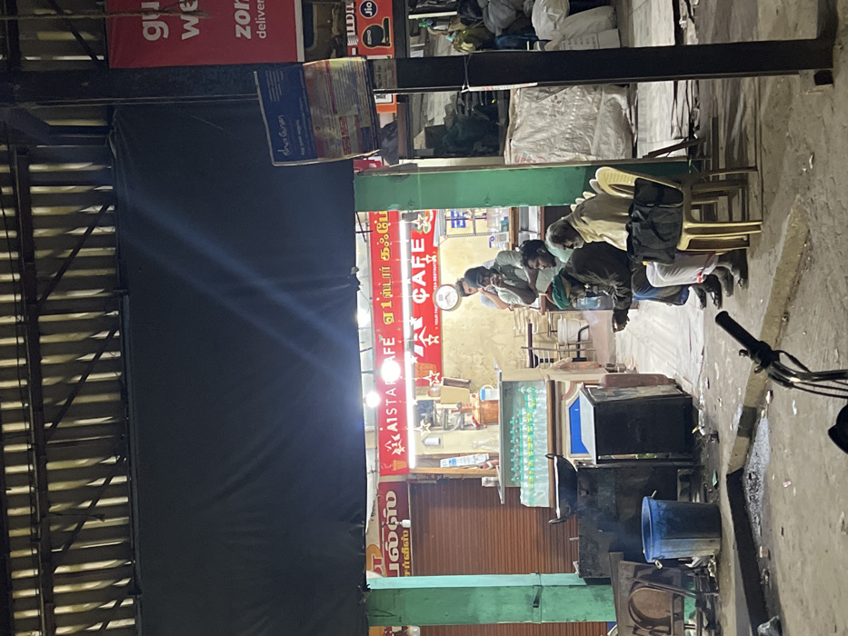 This media file captures a vibrant street scene at night in Palani, India, centered around a brightly lit cafe. The setting is an urban street, possibly an alley or a narrower road, with various commercial establishments.
The main subject is "A1 STAR CAFE," prominently displayed on a red signboard with white text. Below this, the Tamil script "ஏ 1 ஸ்டார் கஃபே" confirms the cafe's name. Inside the cafe, a wall clock is visible, and stacks of bottled water line a counter. Several men are gathered both inside and immediately outside the cafe. A group of men is seated on light-colored plastic chairs facing the cafe, appearing to be patrons socializing or waiting. One man among them wears a dark cap or hood. Another individual is visible behind the counter inside the cafe.
The scene is illuminated by artificial lights emanating from the cafe, casting a glow on the street and its occupants, contrasting with the dark surroundings. To the far left, a large dark tarp or shutter covers what appears to be another establishment or part of a building, with a metallic corrugated roof structure above. On the far right, part of a bicycle's handlebars and front wheel are visible in the foreground. Utility items, including a blue bucket, are placed on the ground near the cafe.
Overhead, several banners are visible. A red banner clearly displays text like "Zor" (likely referring to Zomato) and "delivery," indicating food delivery services. Another banner shows "Jio" and "Idea," advertising telecommunication services. Other visible Tamil text includes "பல்ஸ்" (Pulse) and "CAFE" on the signage, and "Service" and "shalunn" (possibly "salon") on smaller cards. The overall ambiance suggests a casual, bustling evening or late-night gathering spot.