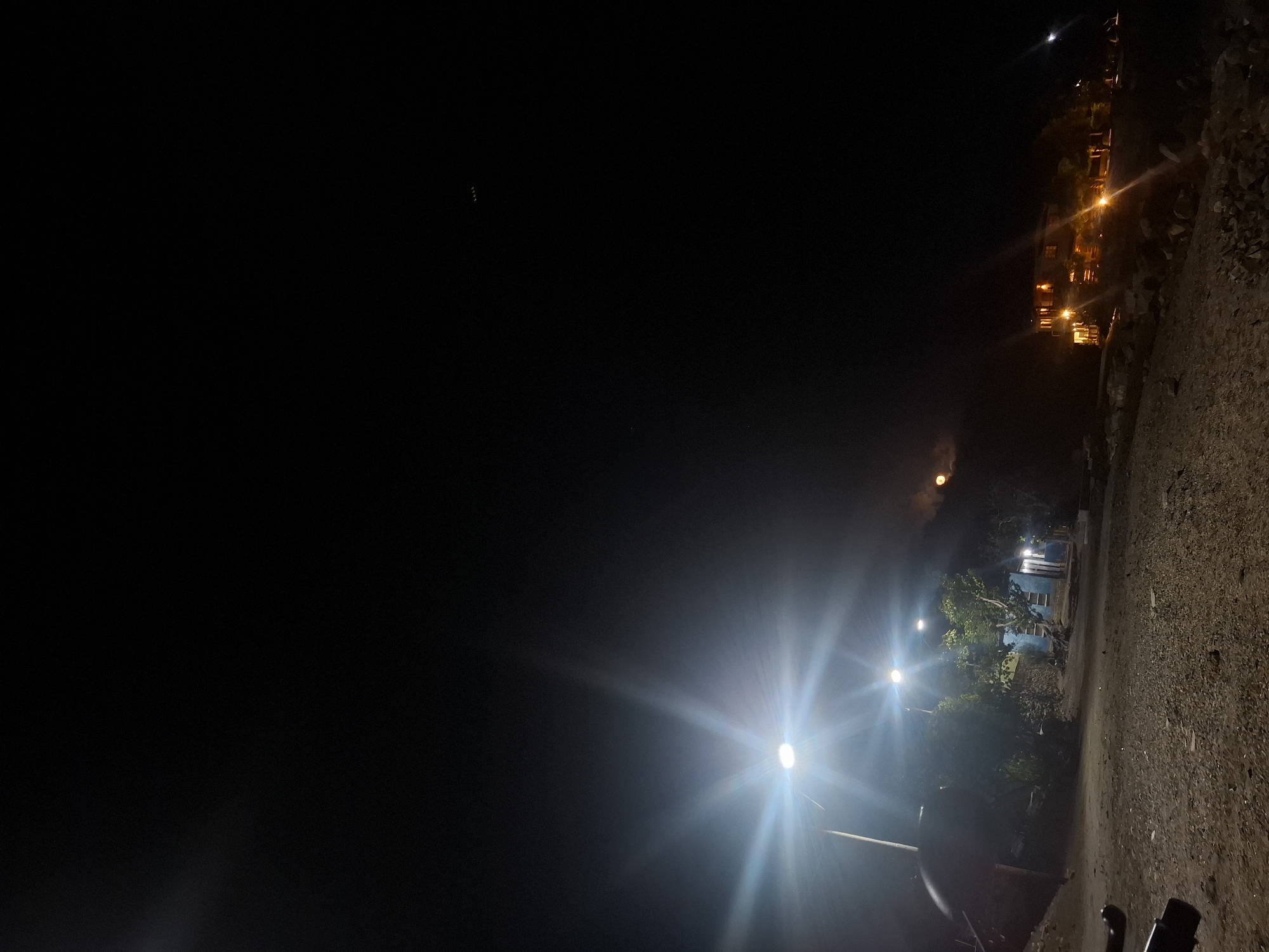 The image depicts a night scene in Pedro González, Venezuela. The foreground shows a dark, gravel-covered area with what appears to be the partial view of a vehicle or motorcycle. Streetlights with bright, starburst-like glows illuminate the scene, interspersed with trees and vegetation. In the distance, a building with lit windows suggests residential or commercial activity. The upper portion of the image is dominated by dark sky, punctuated by a small, bright celestial object, likely the moon or a bright star. A faint, reddish glow is also visible in the mid-ground. The overall impression is of a quiet night in a partially lit area of the city.