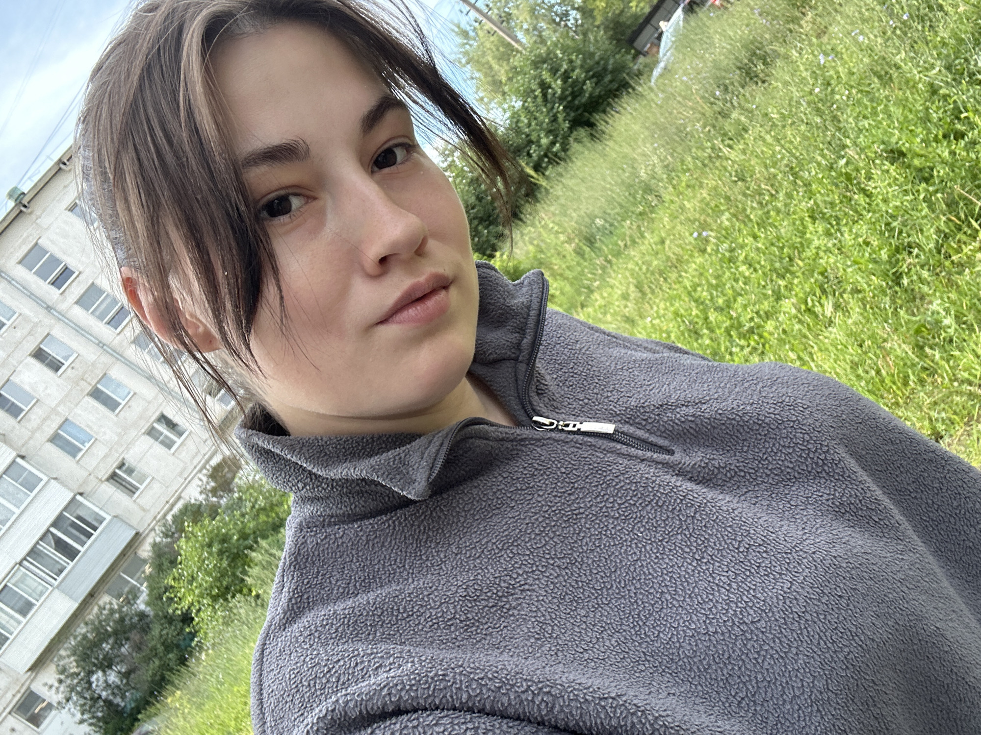 A young woman is taking a selfie outdoors. She is wearing a grey zip-up fleece jacket. Her dark hair is pulled back from her face, with some strands falling forward. She has a neutral expression.
In the background, to the left, is a multi-story concrete building with many windows. To the right, there is lush green grass and trees, indicating an outdoor setting with vegetation. The sky is visible, suggesting daytime. The overall scene appears to be in a residential area with a mix of urban and natural elements. The weather appears pleasant, likely warm given the outdoor setting and the woman's attire. There is no visible text in the image.