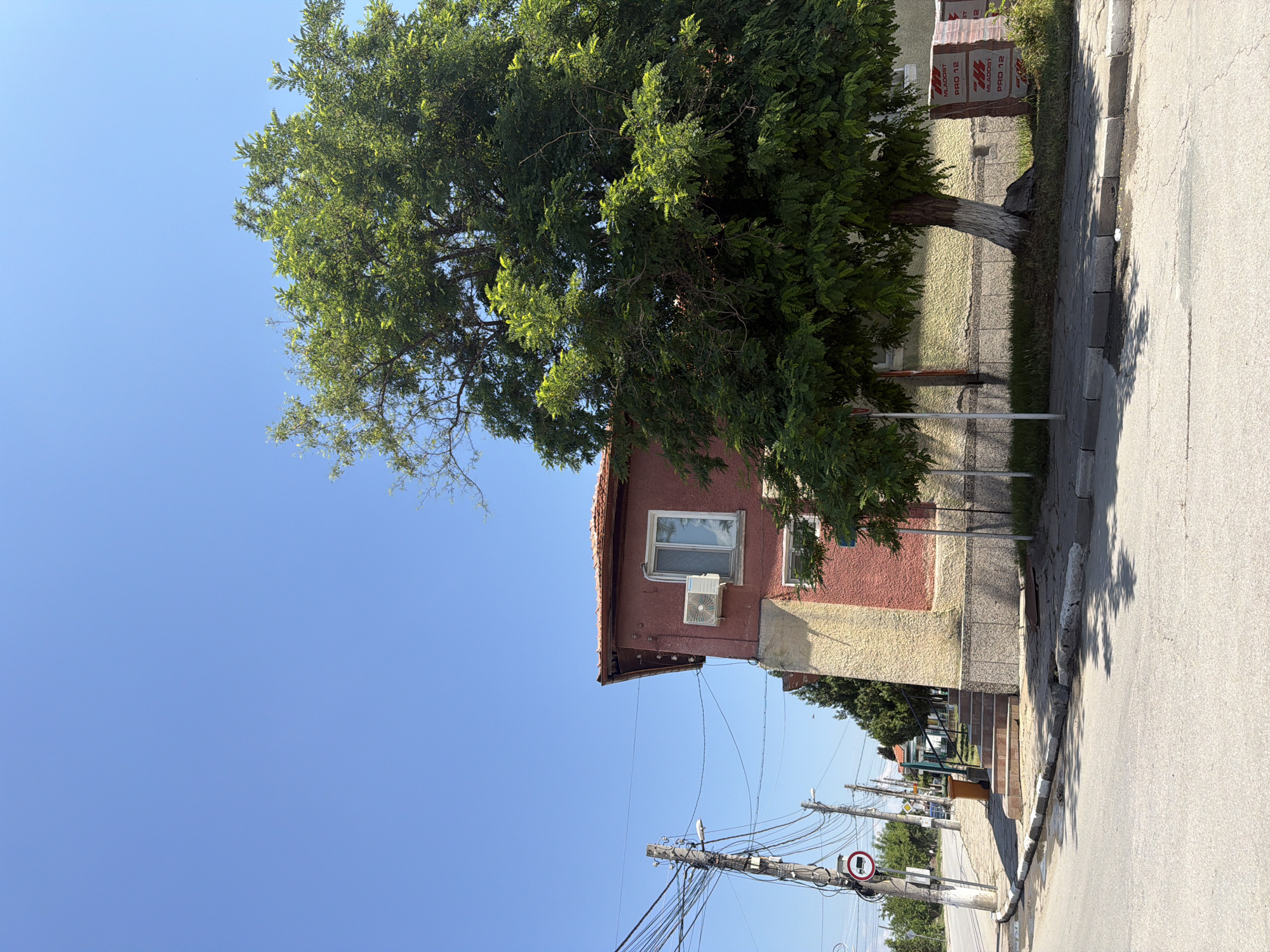 A wide shot captures a sunny day in Zlatitrap, Bulgaria, with a clear blue sky overhead. In the foreground, a paved road curves into the scene. To the left, a sidewalk runs alongside a building with a red facade and a yellow lower section. An air conditioning unit is mounted on the red wall, and a window is visible. Utility poles with numerous wires are positioned near the street. A road sign depicting a truck is also present. A set of stairs leads up to the building's entrance. A large, leafy tree stands beside the building, its branches extending over the sidewalk. Behind the tree, stacks of red-wrapped materials, possibly construction supplies, are visible against the building's wall. In the background, more trees and buildings can be seen, suggesting a residential or semi-urban area. The overall scene is static and undisturbed, with no people or animals in view.