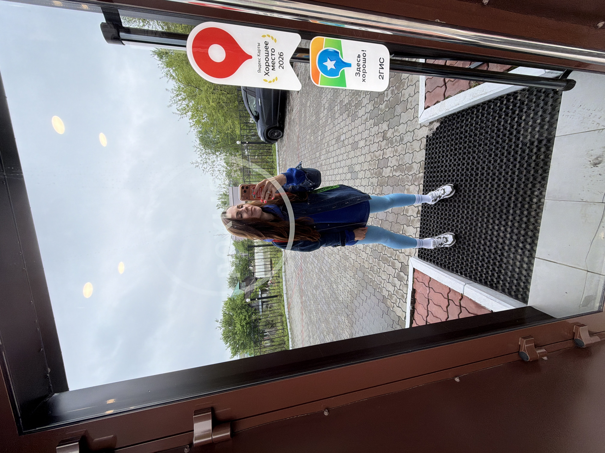 The image shows a reflection in a glass door or window. The reflection features a young woman taking a selfie with a phone. Outside, there is a paved area with a rubber mat near the door. In the background, there's greenery suggesting trees and some buildings behind a fence. Stickers are affixed to the glass, including a red and white one that says "Yandex Maps Good Place 2026" and another with a blue star on it. Based on the stickers (Yandex Maps and 2GIS), it's likely that the location is in Russia. Without additional information, it is impossible to verify the specific location as Birobidzhan.