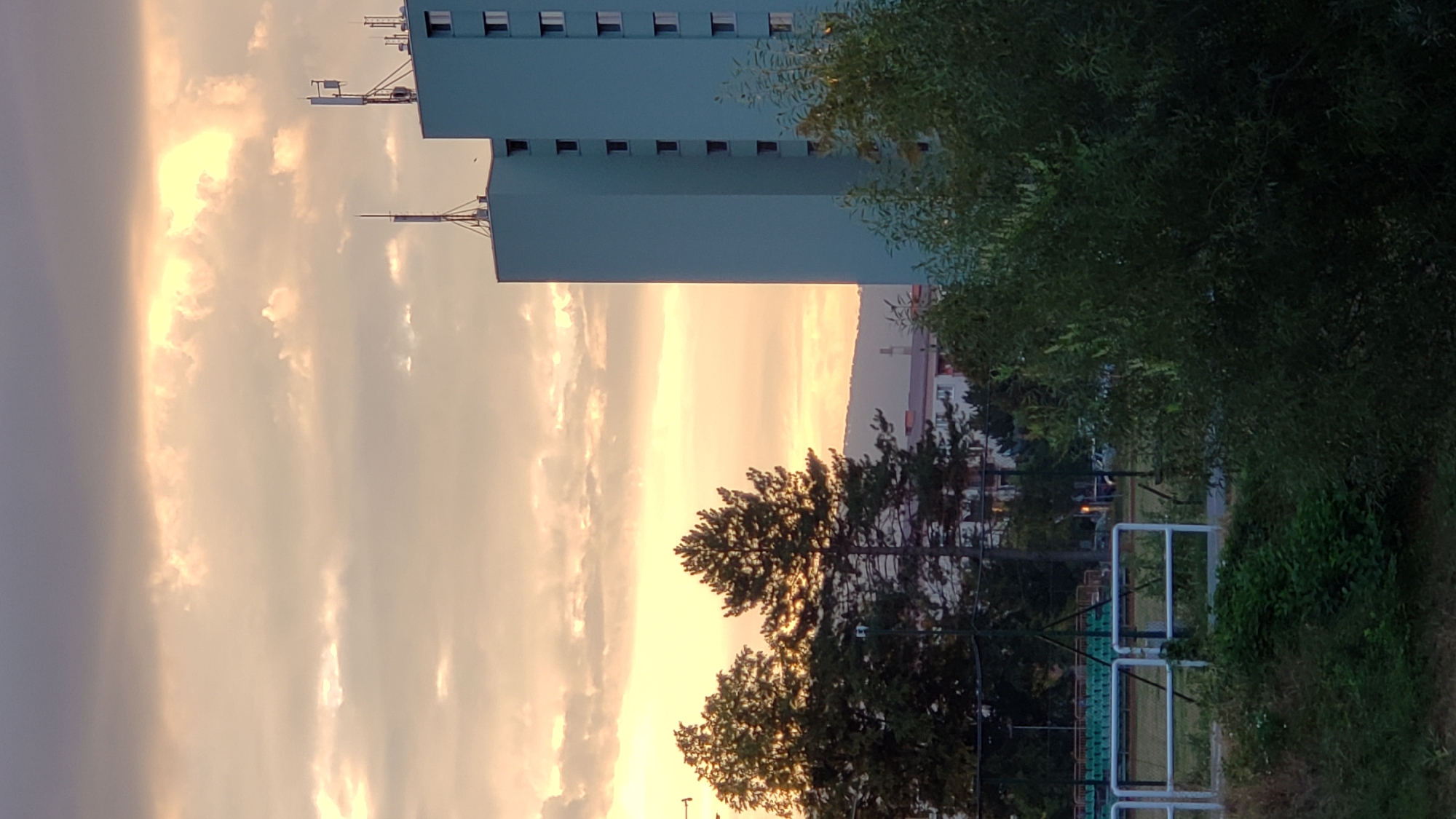 The image shows a residential building in Prešov, Slovakia, at dusk. The sky is filled with dramatic clouds, illuminated by the setting sun, creating a warm orange and yellow glow. Two tall, light blue apartment buildings with numerous windows and communication antennas on top dominate the right side of the frame. In the foreground, lush green trees and bushes partially obscure the view of a sports field with rows of green bleachers. The overall impression is of a quiet, urban landscape at the end of the day.