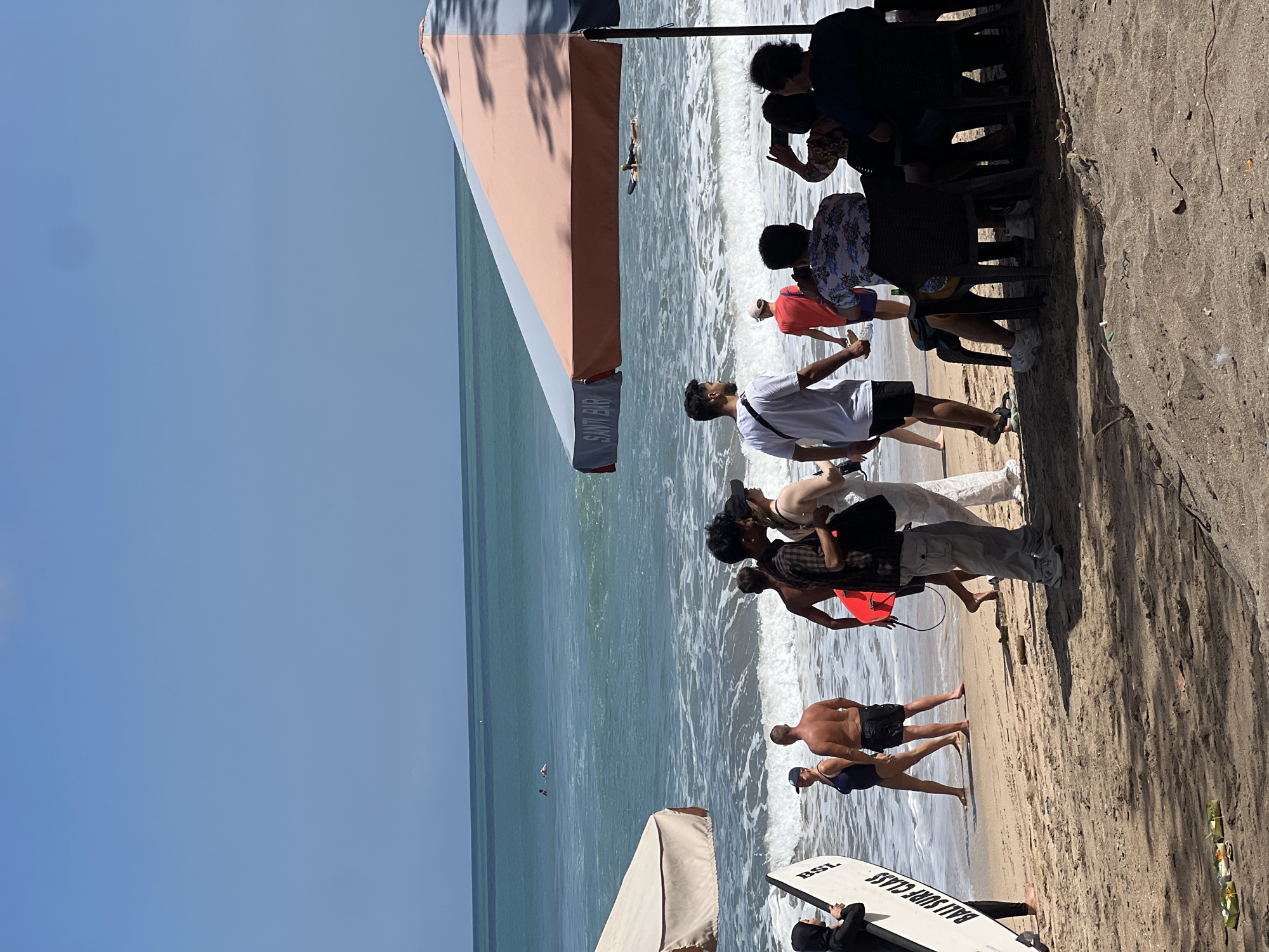 The image depicts a sunny day at Kuta Beach in Bali, Indonesia, with several individuals enjoying the shoreline and the ocean. In the foreground, a person wearing a black hijab and carrying a white surfboard with "BALI SURF CLASS" written on it walks towards the water. To their right, a shirtless man stands looking out at the ocean, with a woman in a blue swimsuit beside him. Several other people are visible further down the beach, some standing and others walking.
The ocean is a vibrant turquoise, with moderate waves breaking on the shore. A few people are in the water, with one individual seen paddling on a surfboard. In the mid-ground, a large umbrella with pink and blue sections shades a group of people seated at tables. The word "SANTI BAR" is visible on the umbrella. The beach itself is sandy, with some scattered debris. The sky above is clear and bright blue. The overall scene suggests a relaxed and active day at a popular tourist destination.