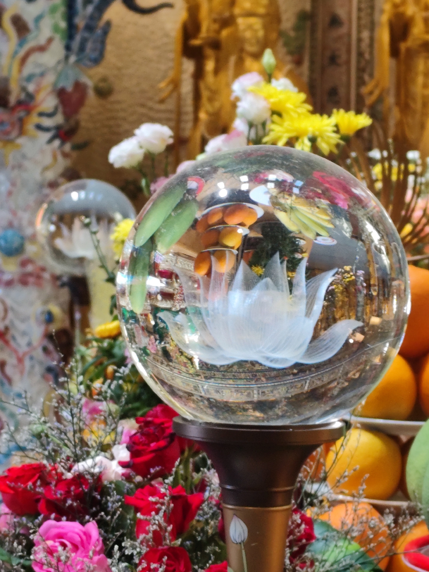 An indoor, eye-level shot focuses on a glass sphere that rests on a dark brown stand. The sphere is filled with reflections of an elaborate Buddhist shrine. A white lotus flower is visible within the sphere, along with other floral and possibly fruit elements. The background of the shrine features gilded statues and colorful, decorative hangings. In front of the sphere, a bouquet of red and pink roses is arranged with delicate, dry greenery. To the right of the sphere, a display of oranges is visible, some whole and some cut. The lighting is soft and diffused, creating a warm and inviting atmosphere. The location is an unknown city.