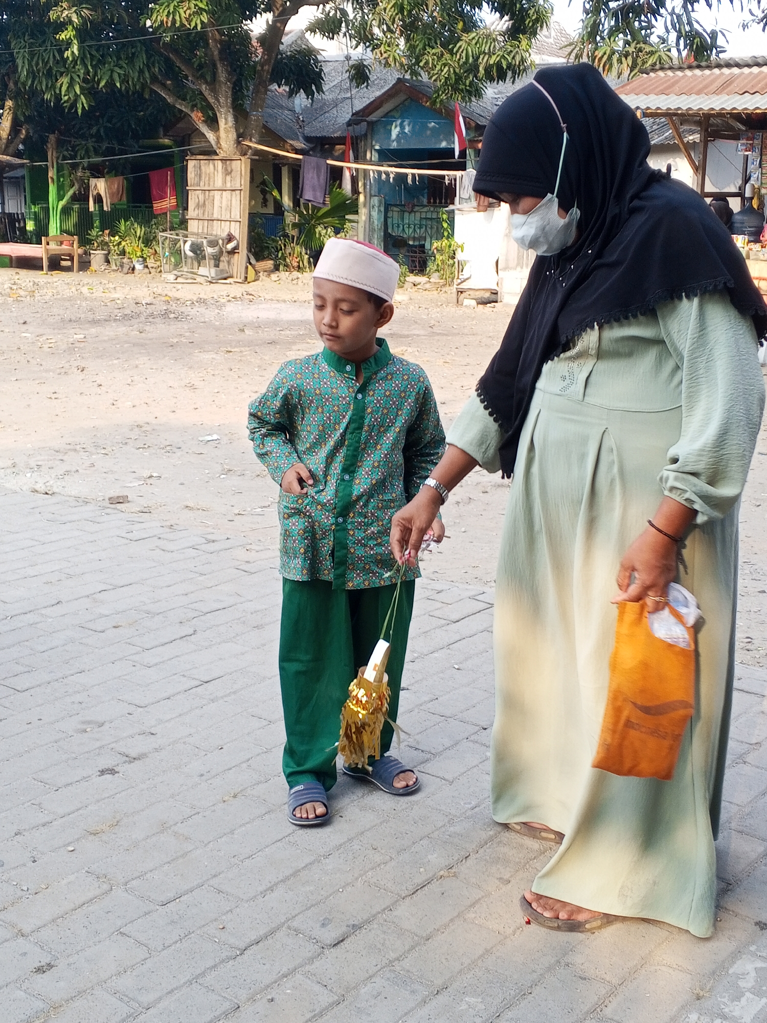 In an outdoor scene on a paved walkway in Ciruas, Indonesia, a young boy and an adult woman are the primary subjects. The boy, positioned on the left, wears a white skullcap, a green shirt with a patterned print, and green pants. He is holding a small, golden, tassel-like object. The woman, on the right, is dressed in a pale green abaya and a black hijab. She is wearing a face mask. She is holding an orange bag in her right hand and reaching out with her left hand towards the boy and the object he is holding. The background shows a residential area with buildings, trees, and laundry hanging. The ground is a mixture of dirt and paving stones.