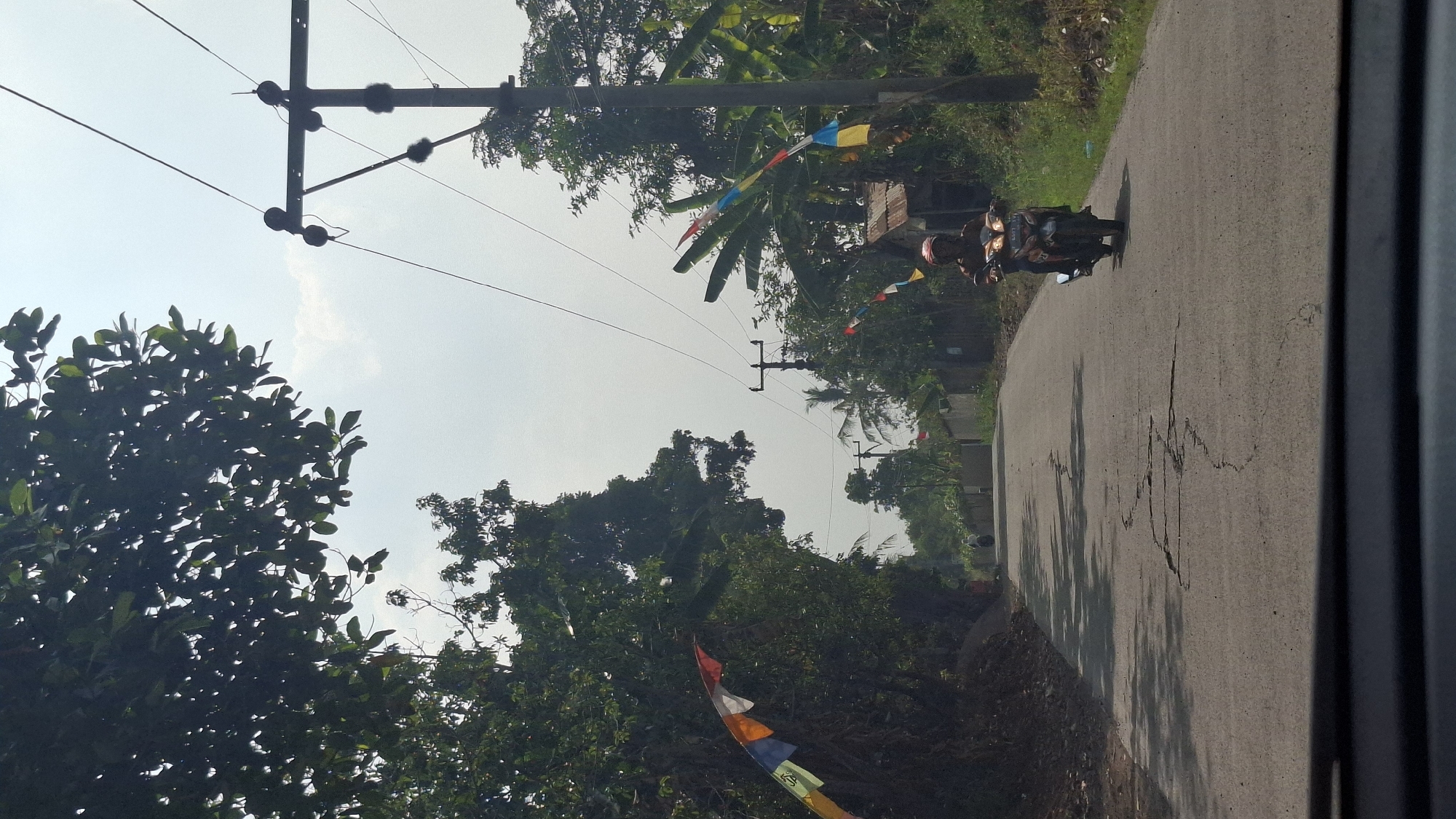 The image depicts a rural road in Sirnagalih, Indonesia, on a bright, sunny day. The road surface is asphalt, showing signs of wear and cracking, with shadows of trees cast across it. On the left side of the road, a dense collection of trees with dark green foliage creates a shaded canopy. On the right, a power pole with electrical wires stands tall, and beyond it, more trees, including what appear to be banana trees, are visible.
Colorful, triangular flags are strung along the left side of the road, attached to the trees. Similar flags are also present on the right side, near the power pole and behind a small, rustic structure.
A single individual is riding a motorcycle down the road, moving away from the viewer. The rider is wearing a helmet and an orange shirt. The motorcycle is dark in color and has its headlights on. In the background, the road continues and appears to lead towards a more built-up area with houses and more trees. The sky is bright and overcast with some wispy clouds. The overall impression is of a typical day on a rural Indonesian road.