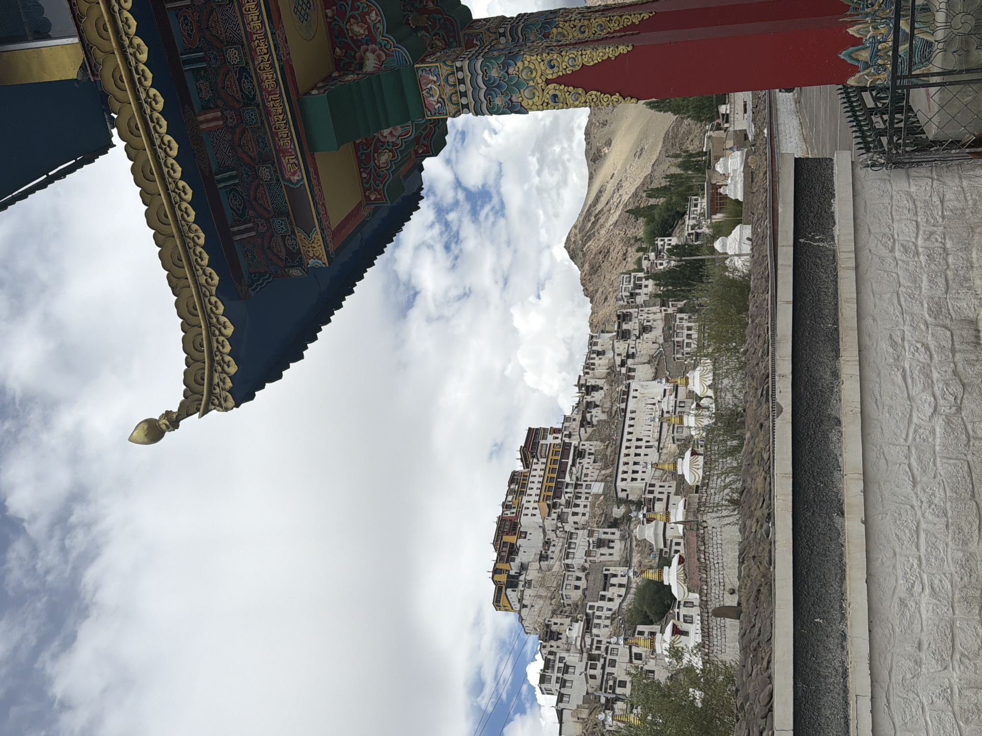 The image depicts the Thiksey Monastery in Thiksey, India. In the foreground, a brightly painted red and gold architectural element, likely a pillar or archway, features intricate colorful patterns and golden decorative trim. This structure partially obscures the view of the monastery complex situated on a hillside in the background.
The monastery itself is a multi-tiered white structure with yellow and red accents, built into the side of a rocky mountain. Numerous white stupas with golden finials are visible in front of the monastery, arranged in rows. The buildings appear to be made of stone and wood, with many windows and flat roofs.
The environment is mountainous and arid, with a pale, dusty mountain rising behind the monastery. Sparse green trees and shrubs dot the landscape. The sky is overcast with dramatic white and grey clouds, suggesting a daytime scene, possibly with a chance of rain. The overall impression is one of a majestic and ancient religious site set against a rugged natural backdrop.