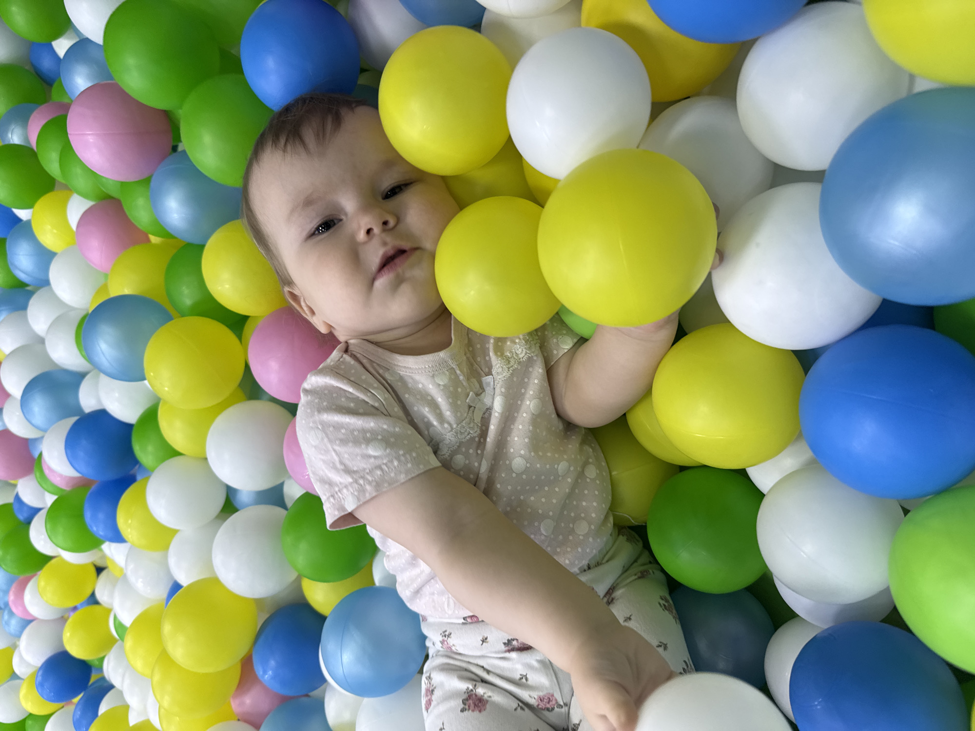 A baby or toddler is depicted lying amidst a vibrant ball pit, surrounded by a multitude of plastic balls in various colors including yellow, white, blue, green, and pink. The child is positioned centrally in the frame, looking directly at the viewer with a somewhat neutral or curious expression.
The baby is wearing a light-colored, short-sleeved top adorned with subtle polka dots and a delicate lace or frill detail across the chest. The pants are also light-colored with a faint floral pattern. The child's right arm is visible, extended into the balls and appearing to hold or interact with several yellow balls.
The ball pit occupies the entire image, from foreground to background, with the balls creating a dense, colorful environment around the child. The scene suggests an indoor play area, likely a children's soft play center or a similar recreational facility. The lighting is even and diffused, consistent with an indoor setting. No specific time of day or external weather conditions are discernible. There is no visible text in the image. The overall context points to a playful moment in Izhevsk, Russia.