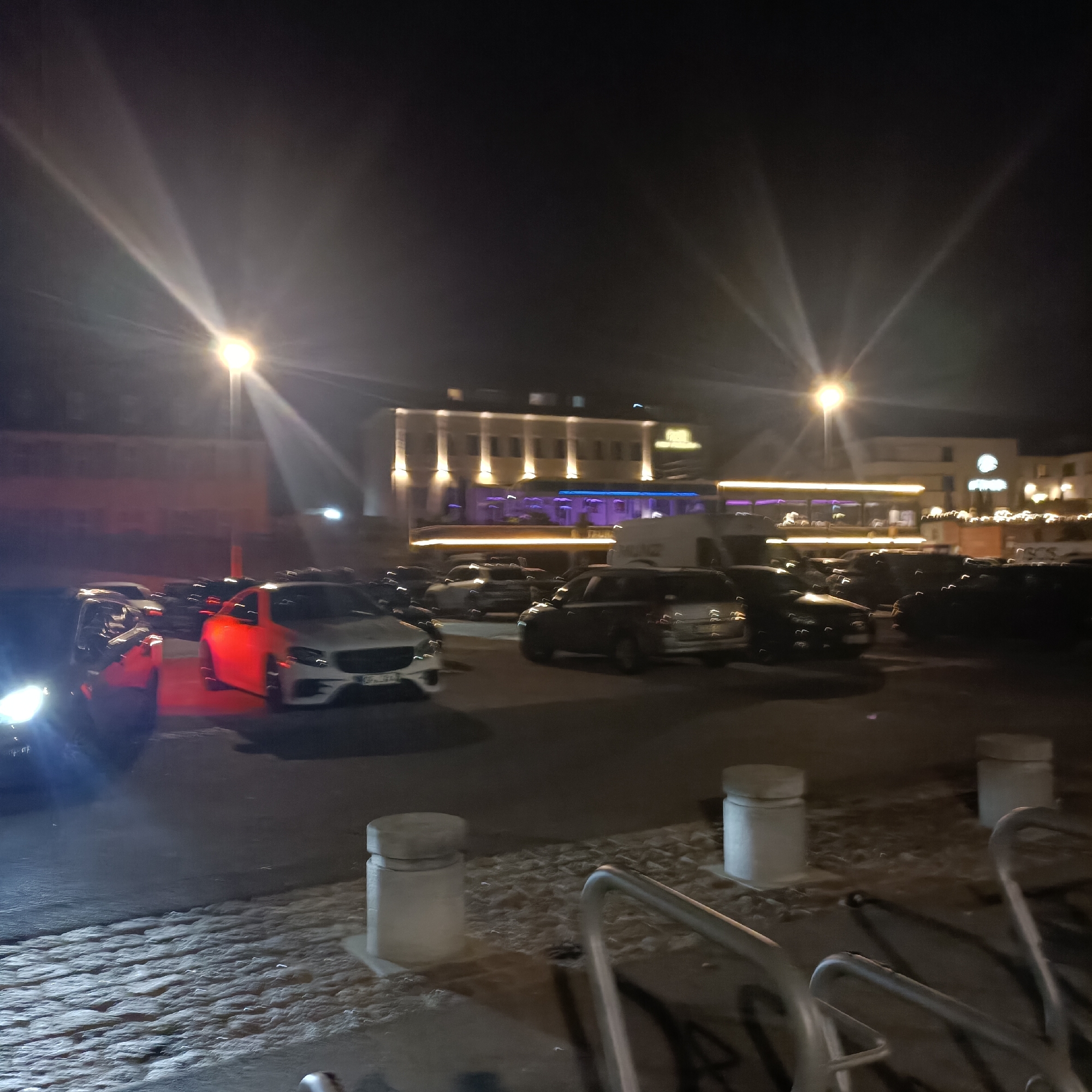The image depicts a nighttime scene in a parking lot, likely in Rüsselsheim am Main, Germany, given the context. Several cars are parked, some with their headlights on, illuminating the scene. In the background, a building with illuminated windows and architectural lighting is visible, suggesting it might be a commercial establishment or residential complex. The ground appears to be paved with cobblestones in the foreground. The lighting conditions create lens flares and a generally dark atmosphere, typical of a nighttime outdoor environment. There are no individuals clearly visible in the image. The scene appears to be a static capture of an empty parking area at night, possibly near a business or entertainment venue.