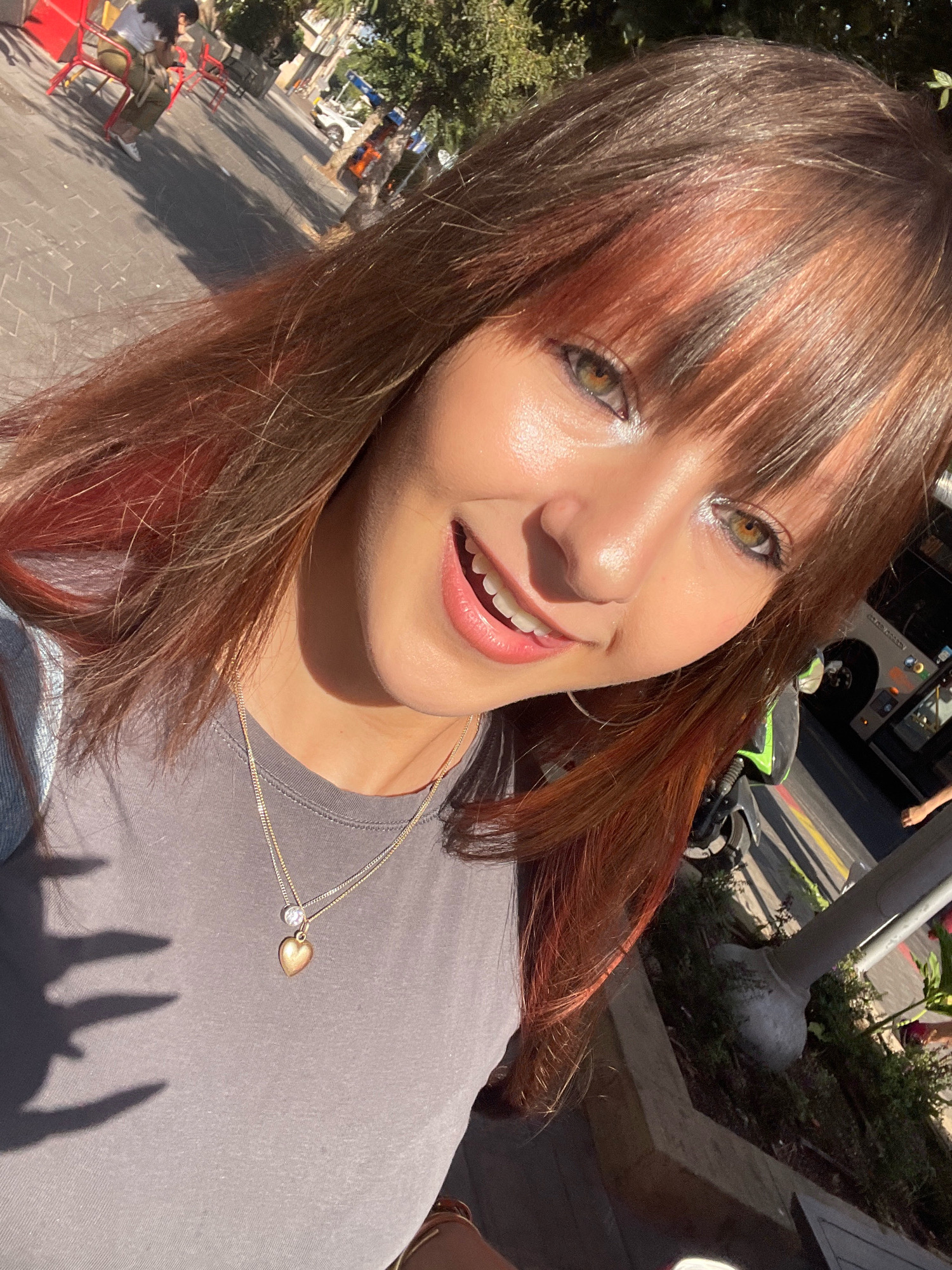 The image is a close-up selfie of a young woman with reddish-brown hair, green eyes, and a smiling expression. She is wearing a gray t-shirt and a gold necklace with a heart pendant. The background appears to be an outdoor urban setting, possibly Raanana, Israel, with a sidewalk, trees, and a bus. Sunlight casts a shadow on the woman's chest, and some greenery and streetlights are visible in the background. The overall tone of the image is bright and casual.