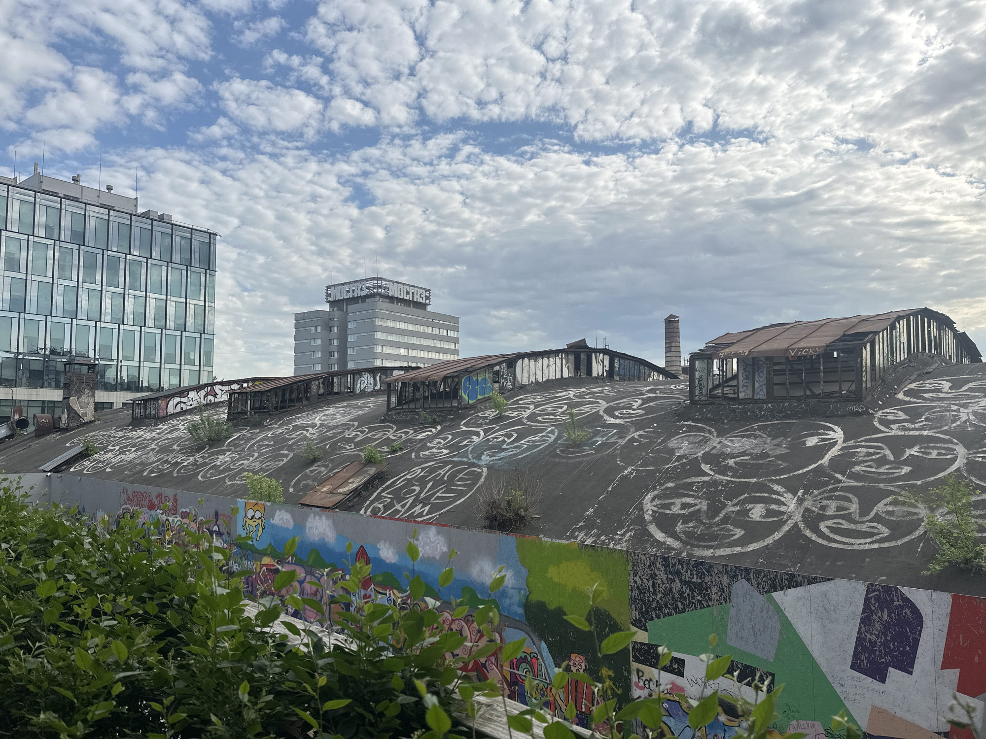 The image depicts a cityscape in Moscow, Russia, showing an old, graffiti-covered industrial building. The building has a curved roof with metal structures on top. The graffiti includes faces, the words "PEACE LOVE FAM", and other abstract designs. A mural is visible on the wall below the roof. In the background, two modern office buildings rise above the industrial structure. The sky is cloudy, with patches of blue. Green foliage obscures the bottom of the image.
