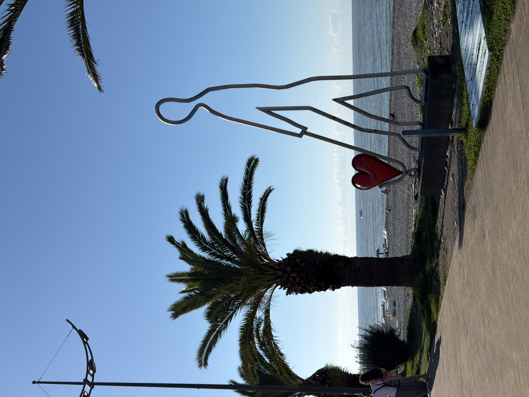 The image depicts an outdoor scene in Batumi, Georgia, likely on the coast. In the foreground, a wooden promenade runs along the left side. Further back, a large, metallic, stylized sculpture of a person is prominently featured, holding a red, heart-shaped object. This sculpture is placed on a concrete base. To the left of the sculpture, two palm trees are visible, with the taller one casting a shadow. Beyond the sculpture, a pebbled beach stretches to the sea, with lounge chairs and umbrellas set up on the shore. The sea is calm, with a boat visible in the distance. A clear blue sky with a few wispy clouds completes the scene. On the left side of the promenade, two individuals are walking away from the camera, one carrying a bag. A street lamp is also visible on the far left of the image.