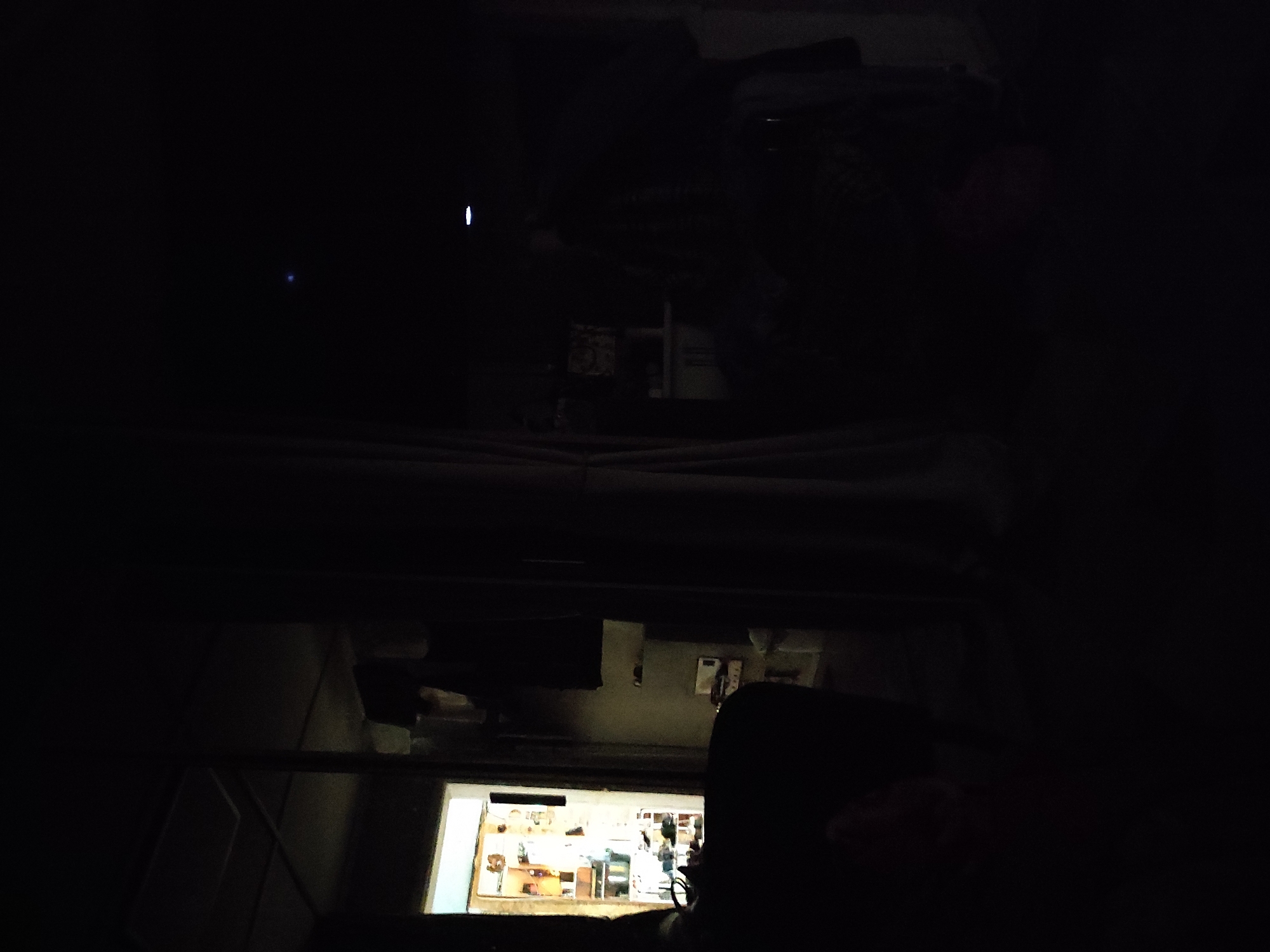 The image depicts a dimly lit interior, likely a room or hallway, with elements suggesting a domestic or storage environment. On the left side of the frame, a doorway or opening leads into a brightly lit space, possibly a kitchen or pantry, filled with shelves stocked with various items. The lighting from this area casts a soft glow on the immediate surroundings. In the foreground and mid-ground, the scene is predominantly dark, with heavy shadows obscuring details. A large, dark, amorphous shape in the lower left corner could be furniture or a person. To the right of the opening, a vertical element, possibly a curtain or partition, divides the space. Further to the right, more darkness prevails, with faint reflections or light sources visible in the upper portion of the frame, including a small blue light and a thin horizontal white light. The overall impression is one of low visibility, with only hints of objects and structures discernible through the darkness. The setting appears to be an indoor environment, with architectural features like a ceiling grid visible in the upper left.