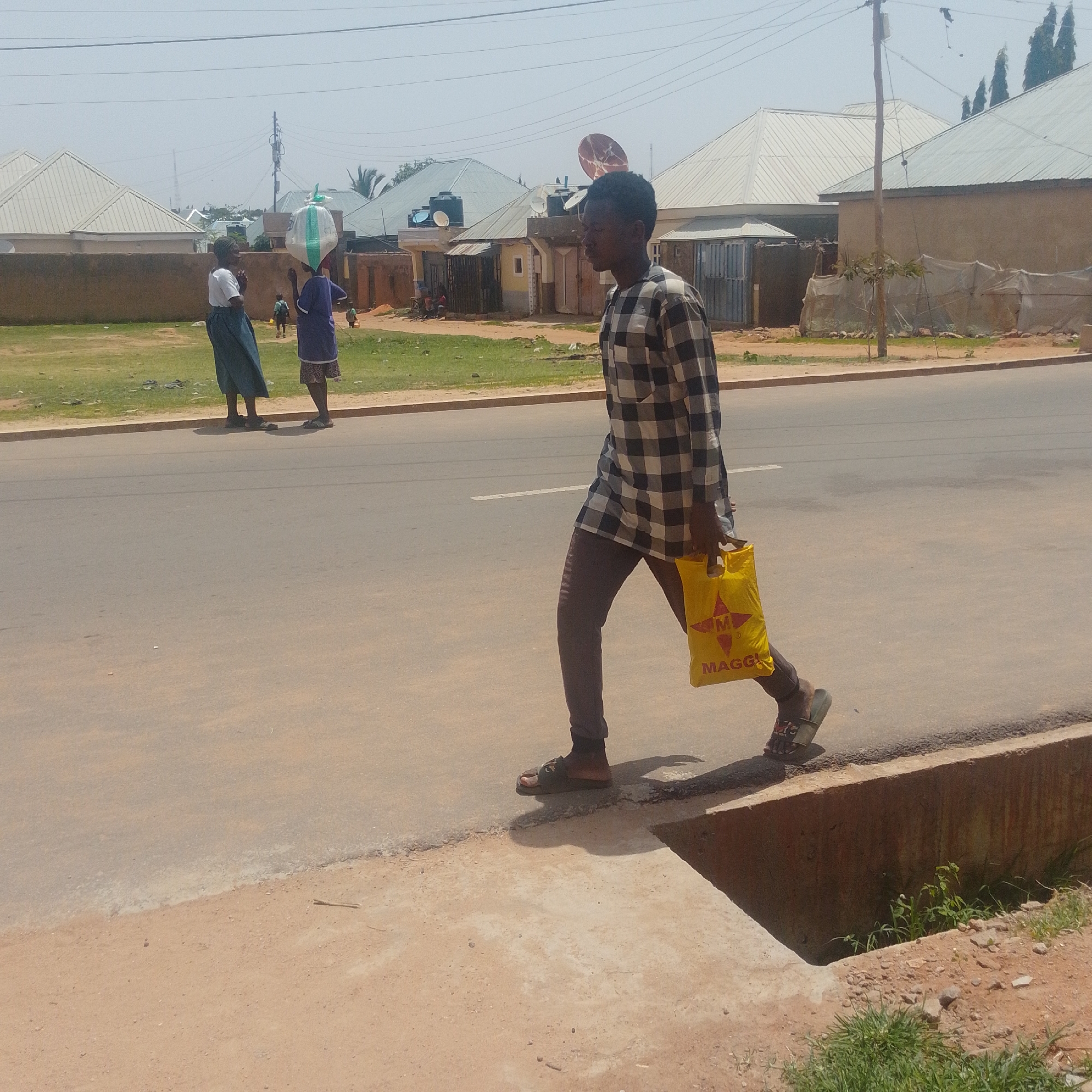 In Gombe, Nigeria, a young man in a checkered shirt and grey pants walks along the side of a paved road. He carries a bright yellow shopping bag with the "MAGGI" logo. To his left, two women are standing on the grassy verge. One woman, wearing a blue skirt and white top, looks towards the camera. The other woman, in a purple top and patterned skirt, has a large white bag balanced on her head. In the background, residential buildings with corrugated metal roofs are visible, along with utility poles and wires. The scene is under a clear, bright sky.
