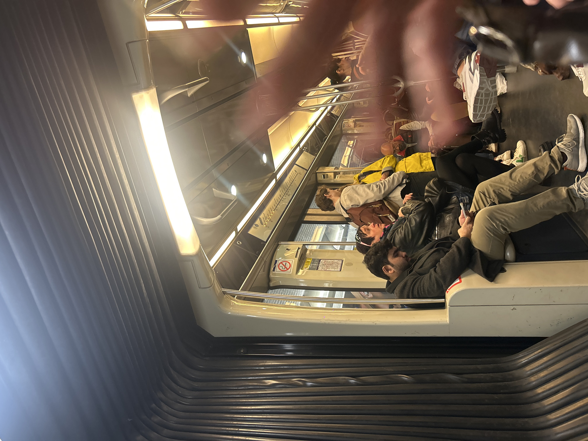 The photo depicts the interior of a subway car, presumably in Paris, France, based on the subway car's design and seating arrangement. Several people are seated, seemingly engaged in various activities, such as looking at their phones and resting. The subway car's design includes horizontal grab rails. The shot appears to be taken from a low angle near the accordion connector between subway cars. A blurry hand is in the foreground, partly obscuring the view.