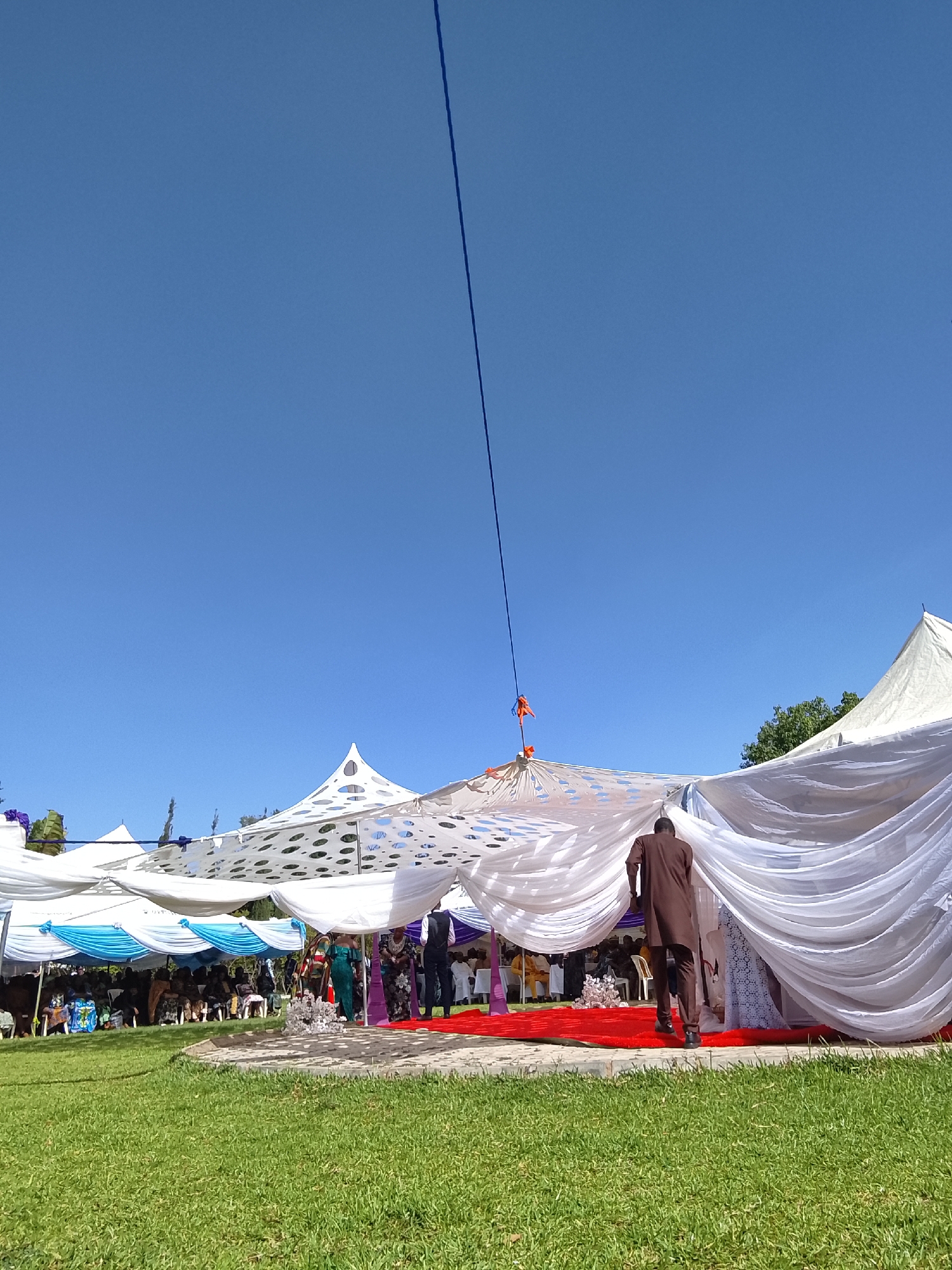 The image shows an outdoor event, likely a wedding, taking place in Unknown, Unknown on a sunny day under a clear blue sky. A large white tent structure is the focal point, decorated with draped white fabric. A red carpet leads into the tent. People are gathered under the tent, seated in rows of chairs. A man in a brown suit is walking towards the tent entrance. There is a green lawn in the foreground. A blue cable is suspended above the tent.