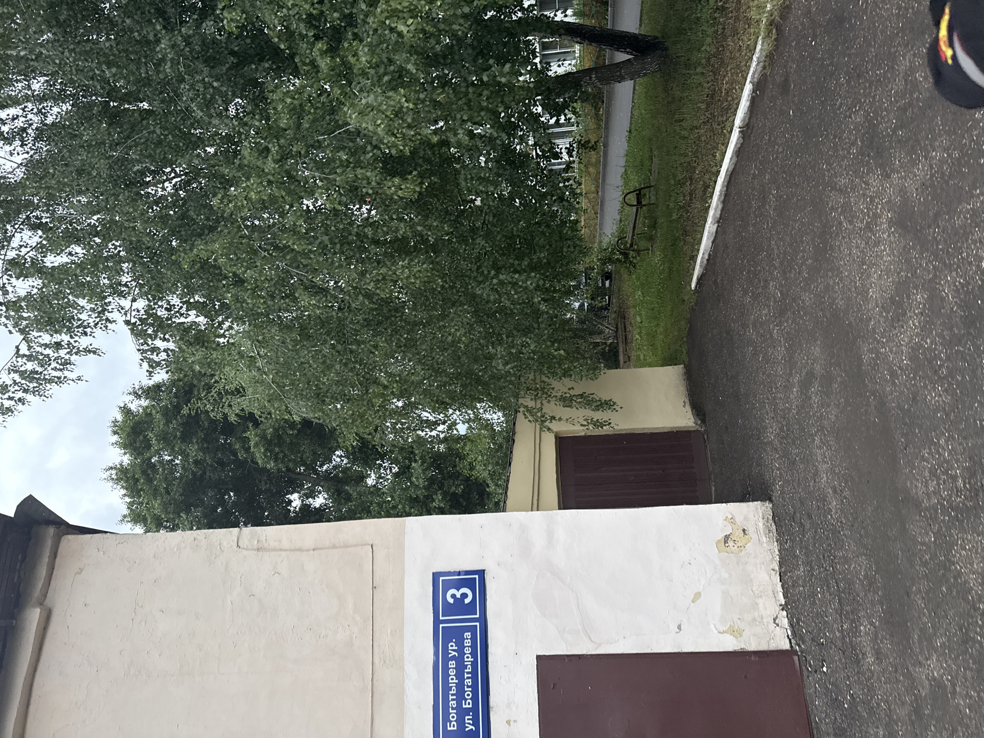 The image shows a street sign with "Богатырев ур." (Bogatirev St.) and "ул. Богатырева" (Bogatirev Street) written on it, along with the number "3". This indicates the location is on Bogatirev Street, number 3. The sign is on the exterior of a pale yellow building. To the left of the building is a dark brown garage door. To the right of the building is a narrow, dark brown garage door and some overgrown greenery. Beyond the building, there are trees and a paved area with a metal bench. The overall environment appears to be an urban residential or service area, possibly a courtyard.