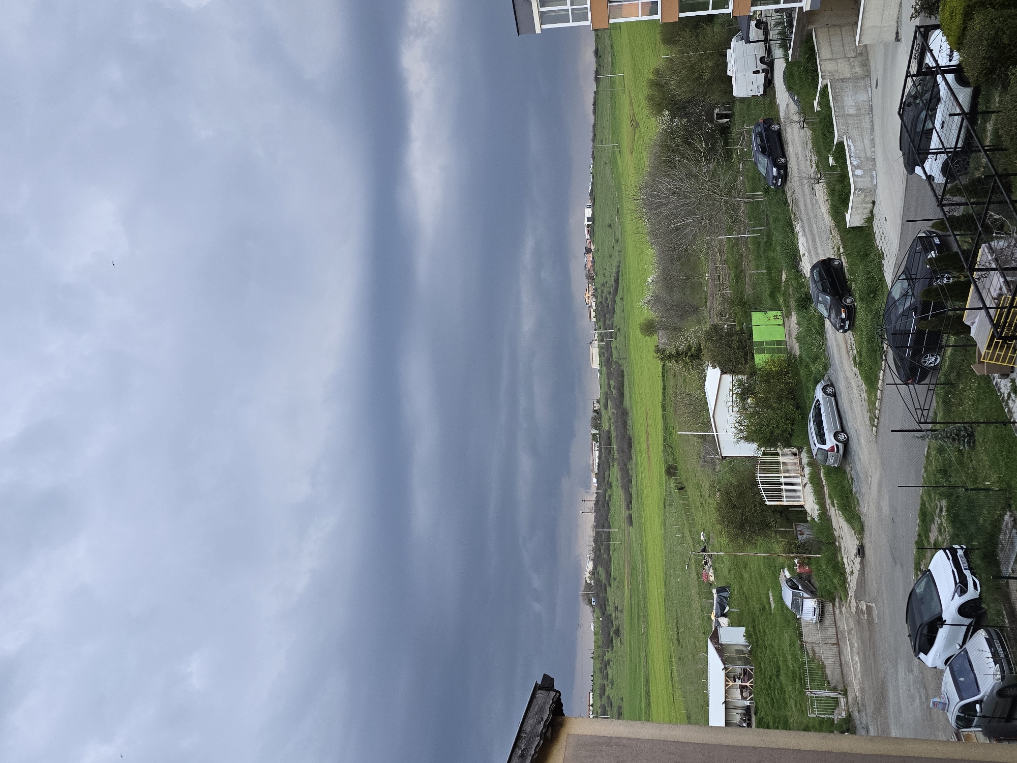 The image presents a panoramic view of a landscape blending residential development with expansive green fields, suggesting an area on the outskirts of Burgas, Bulgaria. The sky occupies a significant portion, featuring a gradient from lighter grey, cloudy conditions on the left to much darker, heavy grey clouds on the right, hinting at dramatic or impending weather. A small bird is visible against the lighter sky.
Below the sky, a vast, vibrant green field stretches towards the horizon, likely agricultural land or pasture. On the right side of the image (when viewed upright), an unpaved road or street runs alongside the field. This road is lined with various parked vehicles, including several dark-colored cars (SUVs or hatchbacks), a white small city car with black detailing, another white car, a silver car, and a white camper van. Visible license plates on some vehicles indicate their origin, with the letter "B" consistent with Burgas.
Parts of modern buildings with windows are visible on the far right, indicating residential structures. Bare trees, green bushes, and grass are scattered throughout the scene. Other visible objects include utility poles extending into the field, a green metal container, and various fences, including a white gate.
The scene depicts a quiet, overcast daytime. There is no specific activity taking place, rather a static portrayal of the environment. The stark contrast between the bright green field and the heavy, dark sky is a notable detail, creating a sense of natural drama. No explicit text is clearly legible beyond the partially visible license plates.