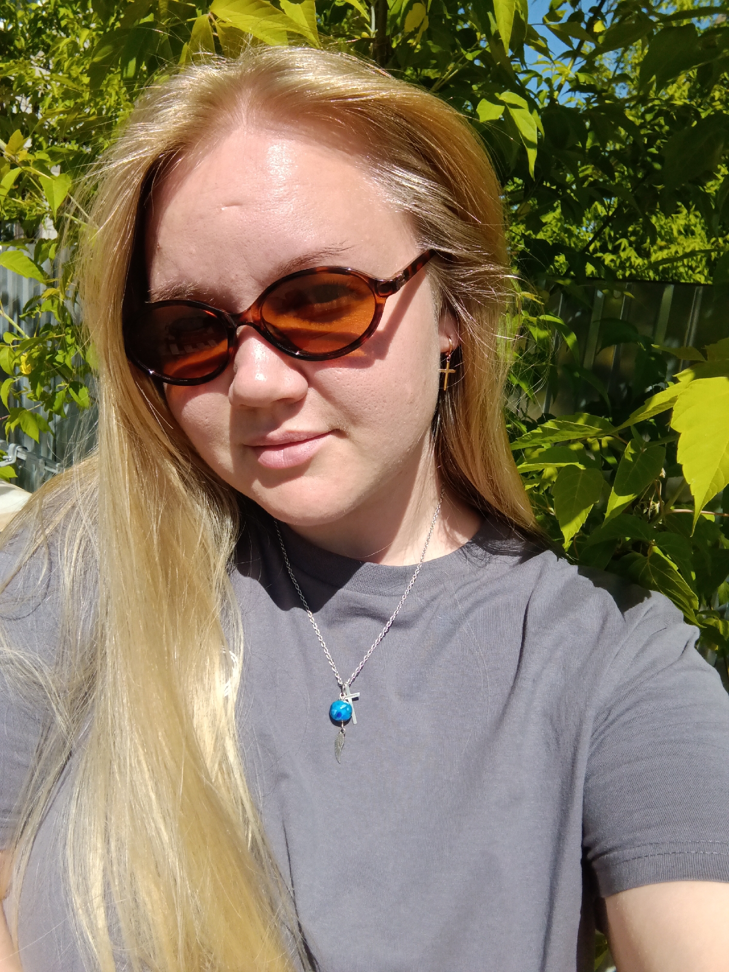 A fair-skinned woman with long, blonde hair poses for a selfie outdoors. She wears brown sunglasses, a grey t-shirt, and a silver necklace with a blue pendant. She has a small gold cross earring in her left ear. Lush green foliage is visible behind her, as well as a glimpse of a silver-colored metal fence. The location is indeterminate, and there is no way to confirm that it is Фоки, Russia from this image.