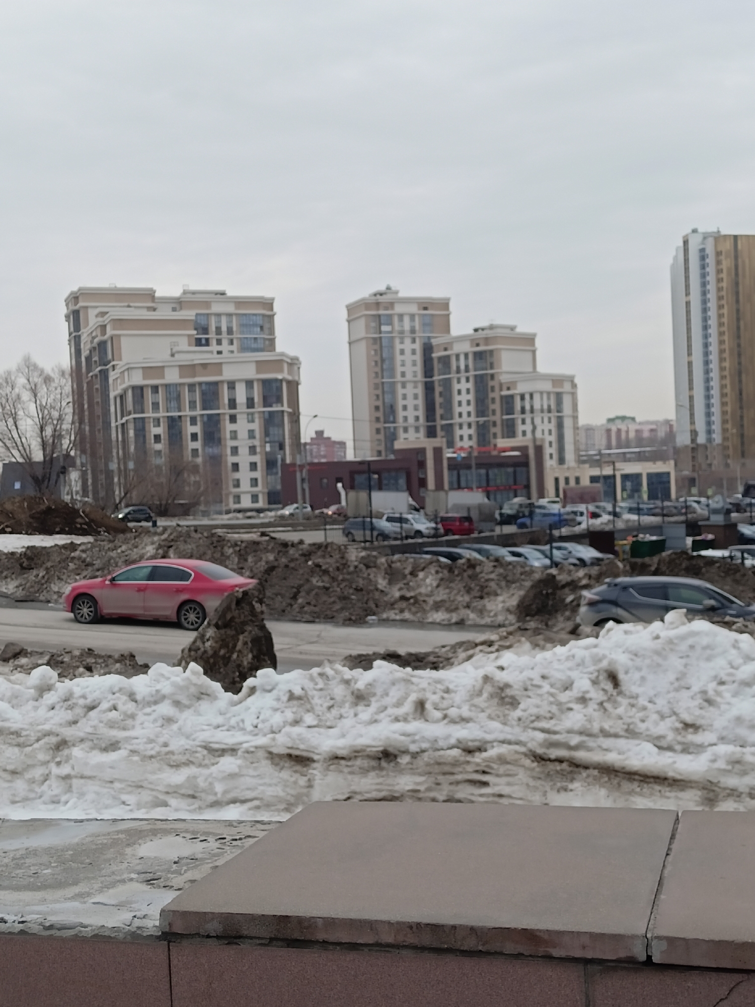 The image depicts a street scene in what appears to be a city, likely in Novosibirsk, Russia. The foreground is dominated by piles of dirty snow and a concrete ledge. In the middle ground, there's a road with cars, including a red sedan, and a parking lot filled with vehicles. Behind the parking lot are multi-story buildings, including one tall skyscraper and several mid-rise apartment complexes. The sky is overcast, suggesting a gray, wintery day. The overall environment suggests a post-snowstorm cleanup in an urban area.