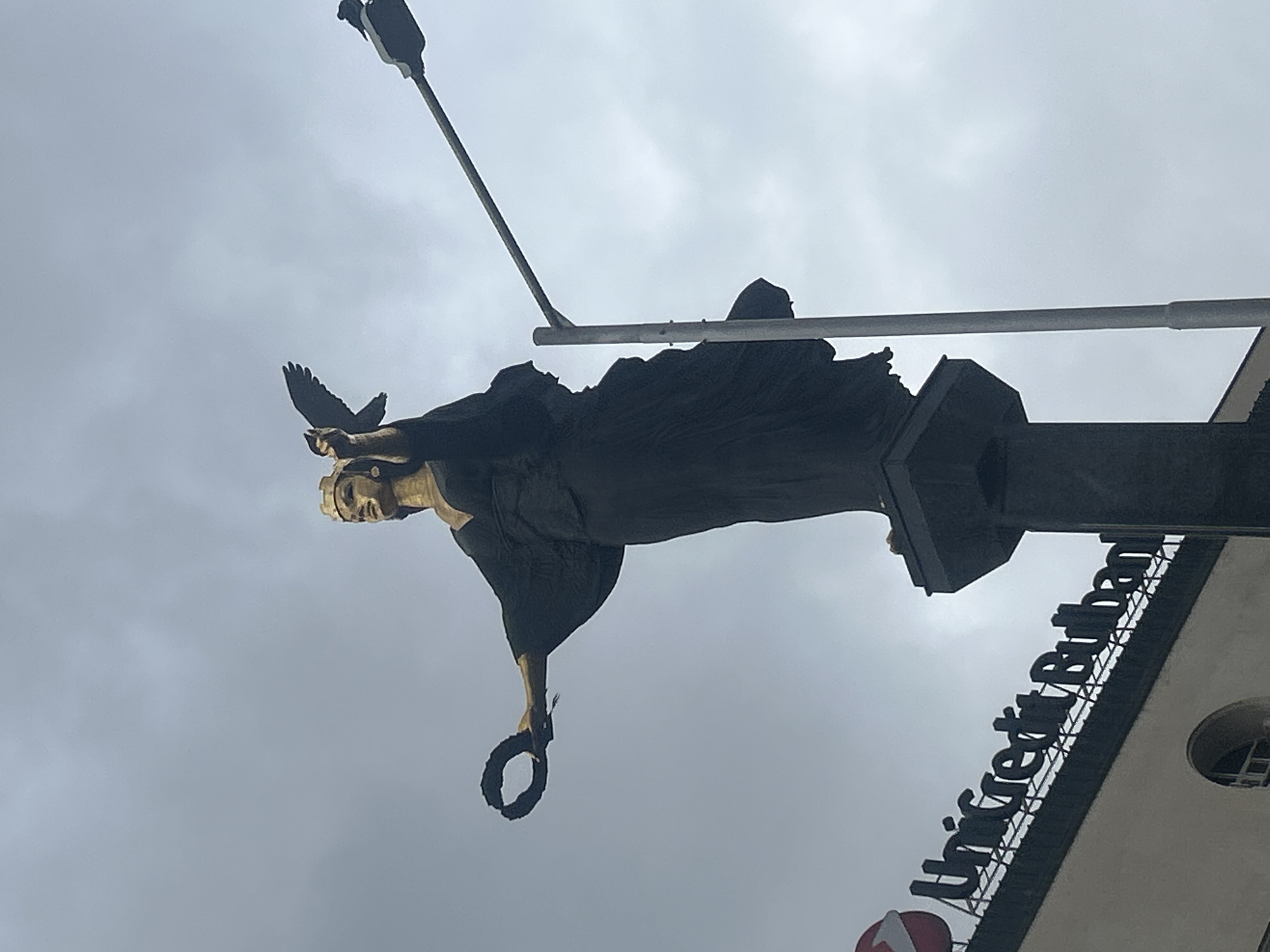 A low-angle shot of the Monument to the Soviet Army in Sofia, Bulgaria. The bronze figure of a woman stands atop a pedestal, holding a wreath in one hand and a dove in the other. A street lamp and building are visible in the background.