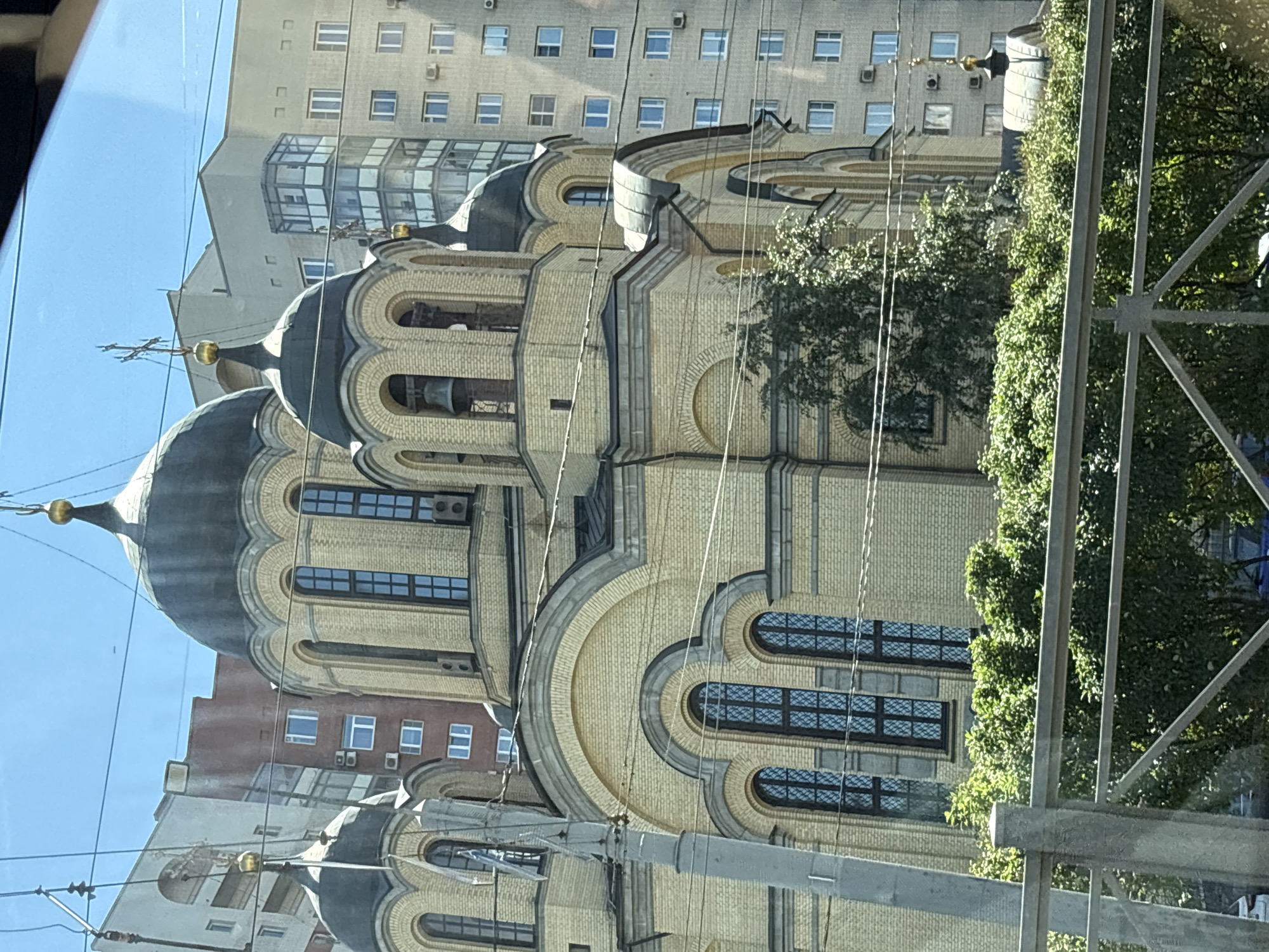 The image captures an outdoor daytime scene in Molodtsovo, Russia. The central focus is a large, pale yellow brick building, likely a church, with multiple dark domes topped with crosses. The building features arched windows with dark panes and decorative brickwork. To the left of the church, a tall, gray concrete utility pole is visible. Behind the church and to the right, several modern apartment buildings with numerous windows are present. Lush green trees and foliage are in the foreground, partially obscuring the lower section of the church. A metal fence runs horizontally across the bottom of the image. The sky is clear and blue.