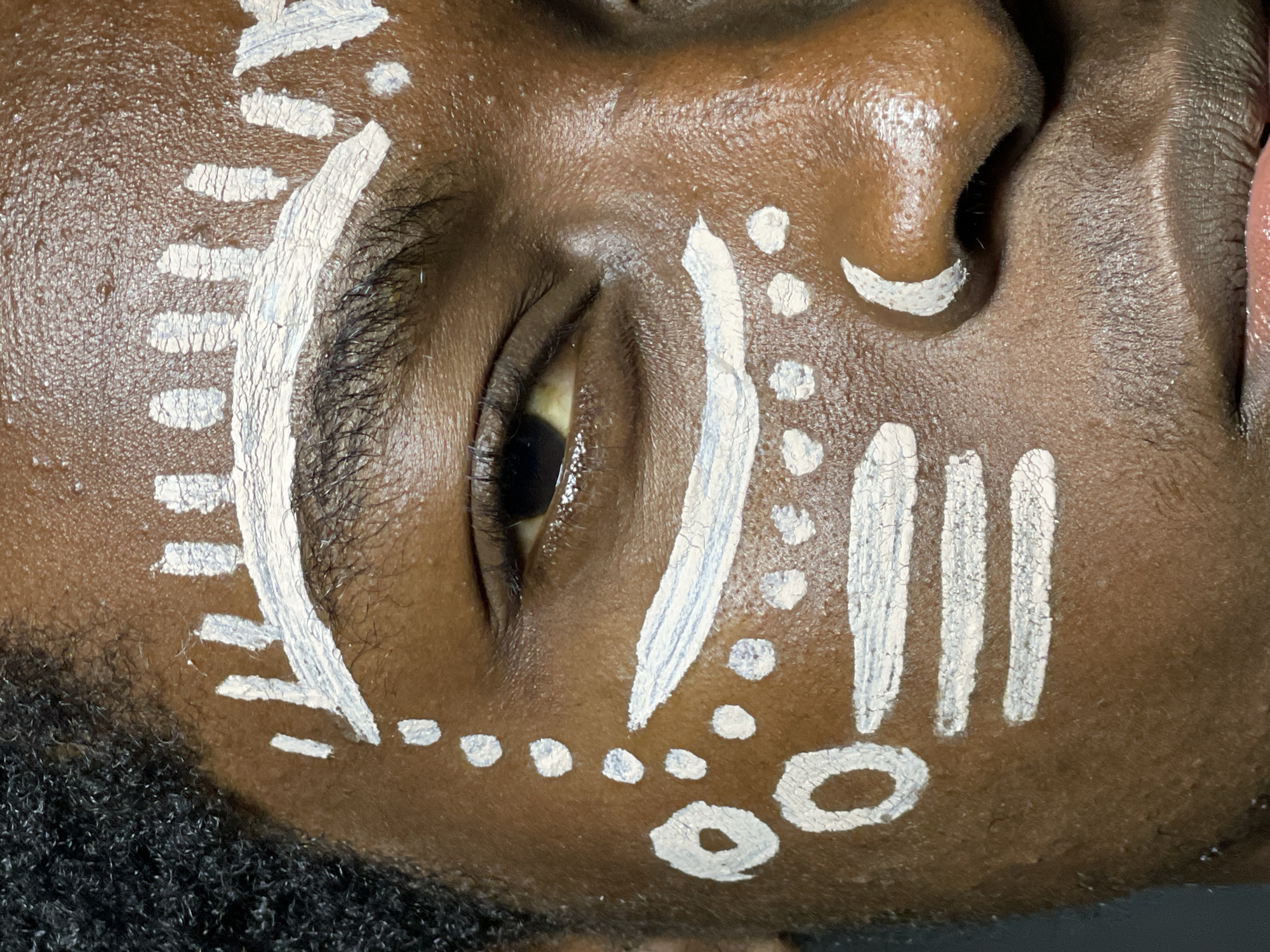 This is a close-up, eye-level, full-frame shot of the face of a person of African descent, adorned with white face paint. The face paint features a series of geometric patterns and dots, applied in a style that evokes traditional African markings. A thick, white line arches above the left eyebrow, with smaller vertical strokes extending upwards from it. Below the eye, a line of white dots runs down the cheek, interspersed with larger circular and rectangular shapes. The skin has a rich, dark complexion and appears slightly oily, with visible pores. The individual's hair, visible on the left side of the frame, is dark and tightly curled. The eye is open and looking slightly to the left, revealing a hazel iris. The lighting is soft and focused on the face, highlighting the texture of the skin and the application of the paint. The background is dark and out of focus, providing no contextual information about the location. The image appears to have been taken indoors, possibly in a studio setting. There are no other people or objects clearly visible in the frame. The overall impression is one of artistic expression and cultural representation through body art.