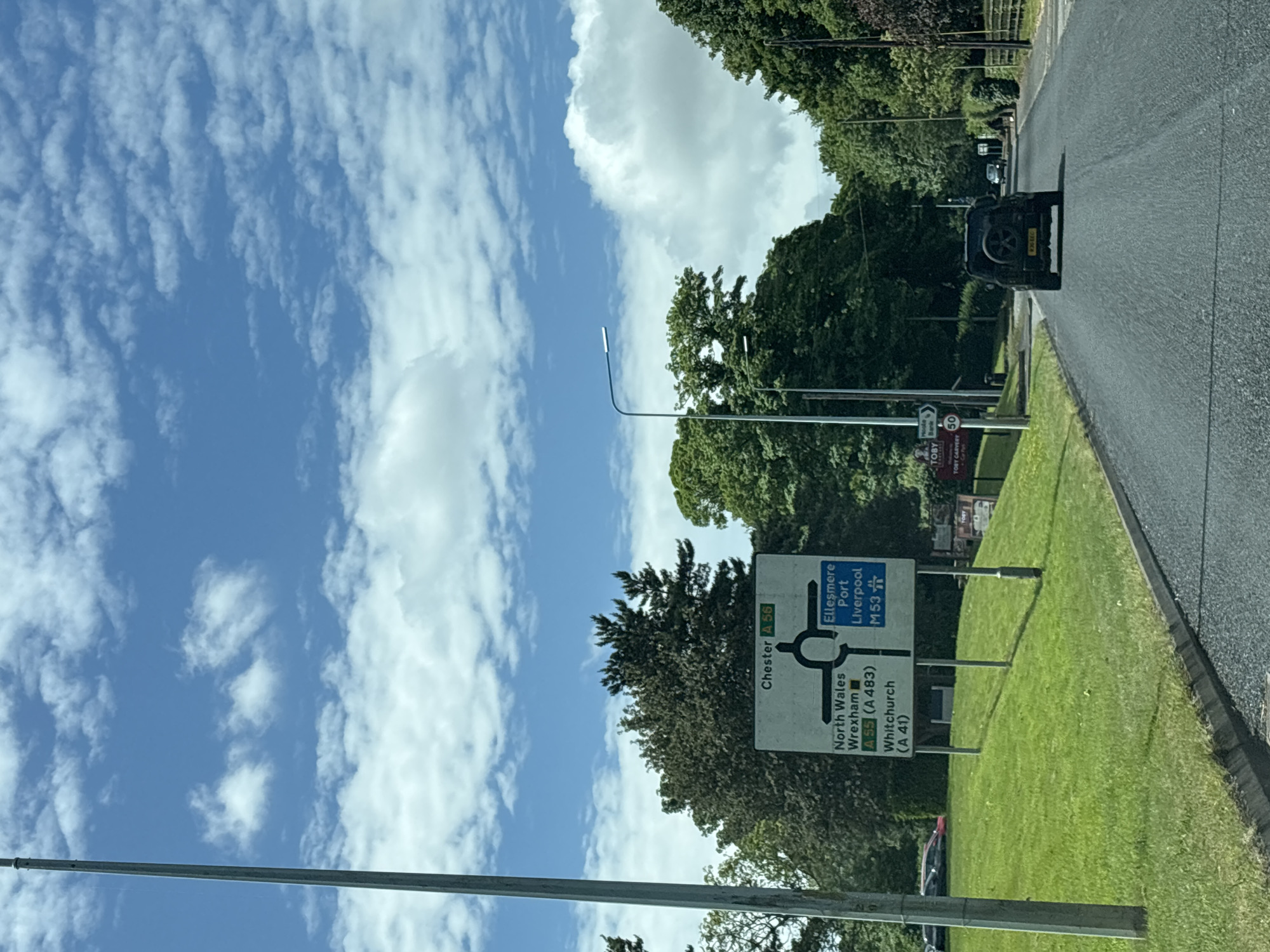 The image depicts a road scene on a sunny day, with a blue sky filled with scattered clouds. The focus is on a road sign indicating directions to various locations. The sign is predominantly white with black lettering. It shows directions to Chester (A 56), North Wales, Wrexham (A55/A483), Whitchurch (A 41), Ellesmere Port, Liverpool, and the M53. The sign is located beside a grassy verge and trees. There's a black SUV vehicle visible on the road in the background. Another sign is also visible indicating a "TOBY CARVERY 50".