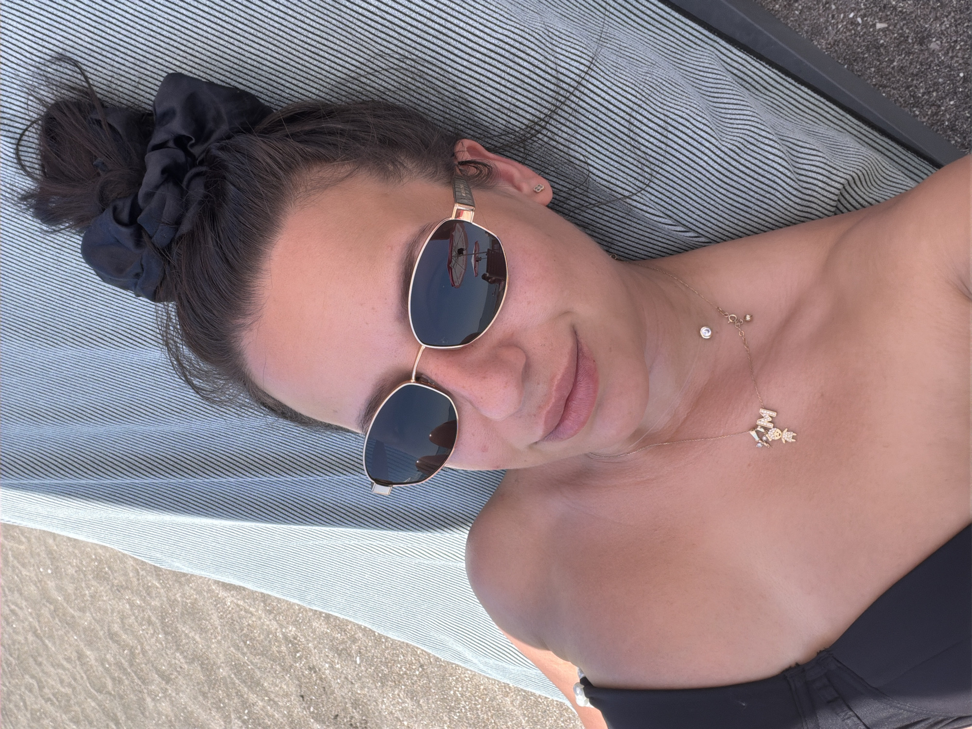 A woman lies on a striped lounge chair on a beach. She is wearing a black bikini top, sunglasses, and a necklace with a teddy bear charm and the letter "M". Her hair is pulled up in a messy bun with a black scrunchie. The beach appears to be sandy with some small pebbles. The background shows a body of water and a blurred reflection of beach umbrellas in the sunglasses. The location is identified as Burgas, Bulgaria.