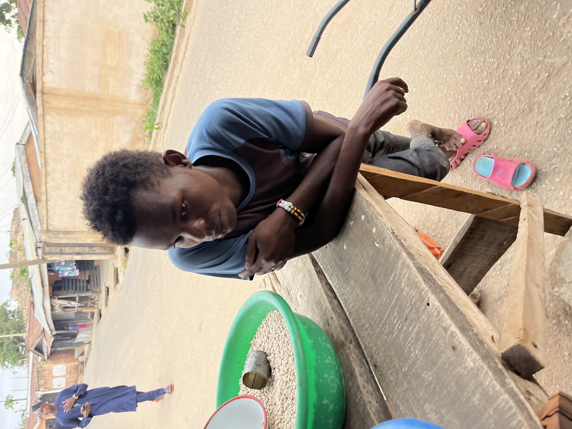 In Jos, Nigeria, a young man sits at a weathered wooden stall on a street. He is resting his arms on the stall, near a green bowl filled with what appears to be grains and a metal scoop. He wears a blue shirt, dark pants, and a colorful beaded bracelet. Next to him are pink and blue sandals on the ground. In the background, a man in a blue outfit walks on the side of the road in the direction away from the camera. The setting includes buildings and street structures indicative of an urban environment.