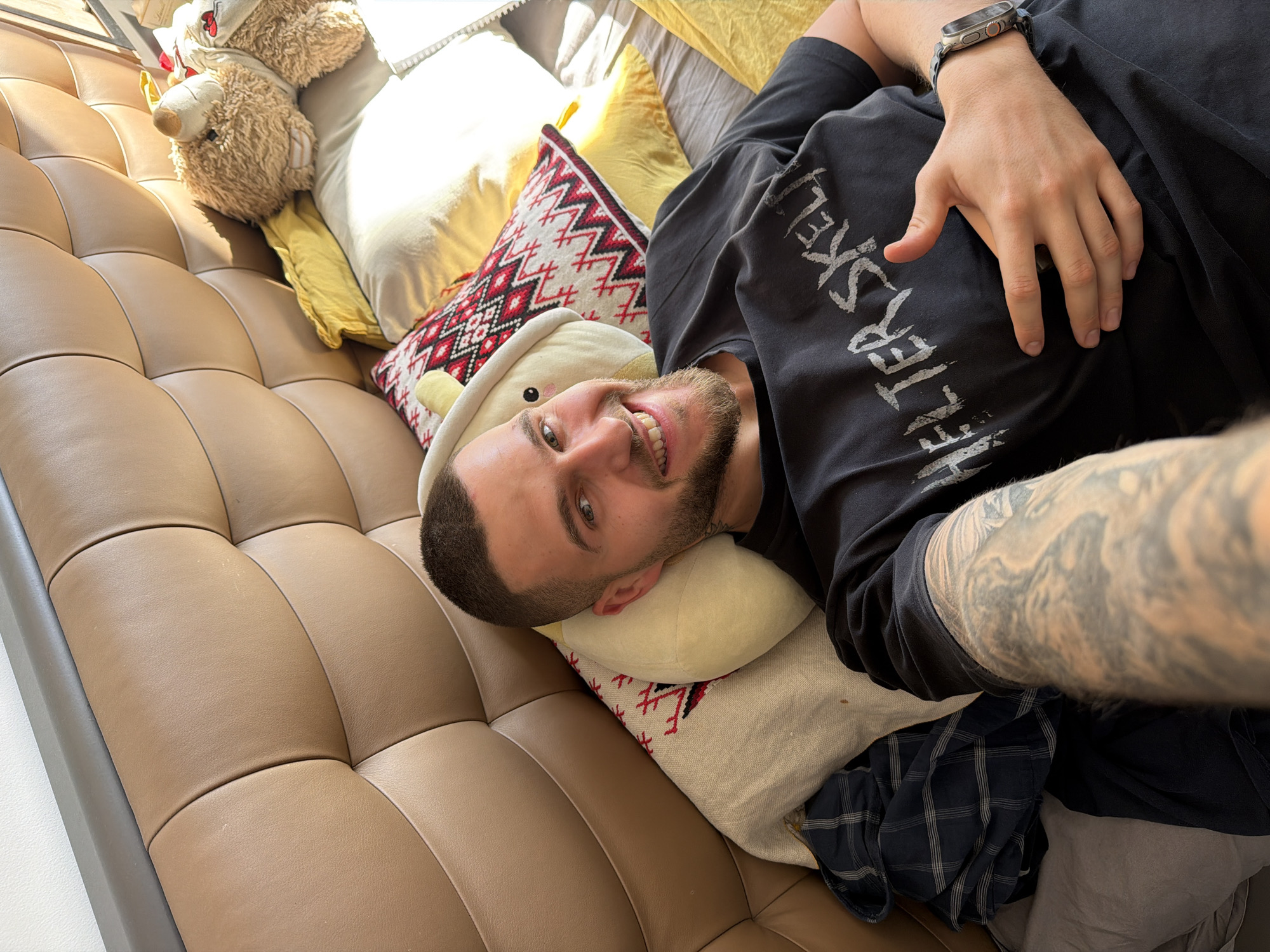 A selfie taken from a high angle shows a man lying on a tufted, brown leather sofa. He is positioned with his head on a small, stuffed, animal-shaped pillow that is on top of a patterned, square pillow. A grey or light-colored blanket is under the patterned pillow. To the left are two stuffed teddy bears. A yellow pillow is behind them.
The man has a short haircut and a beard. He is smiling. He is wearing a black T-shirt with white writing on it. He is also wearing a watch. A tattoo is visible on his forearm. The scene is indoors. This image does not contain information that would permit a determination of location in the city of Sofia, Bulgaria.