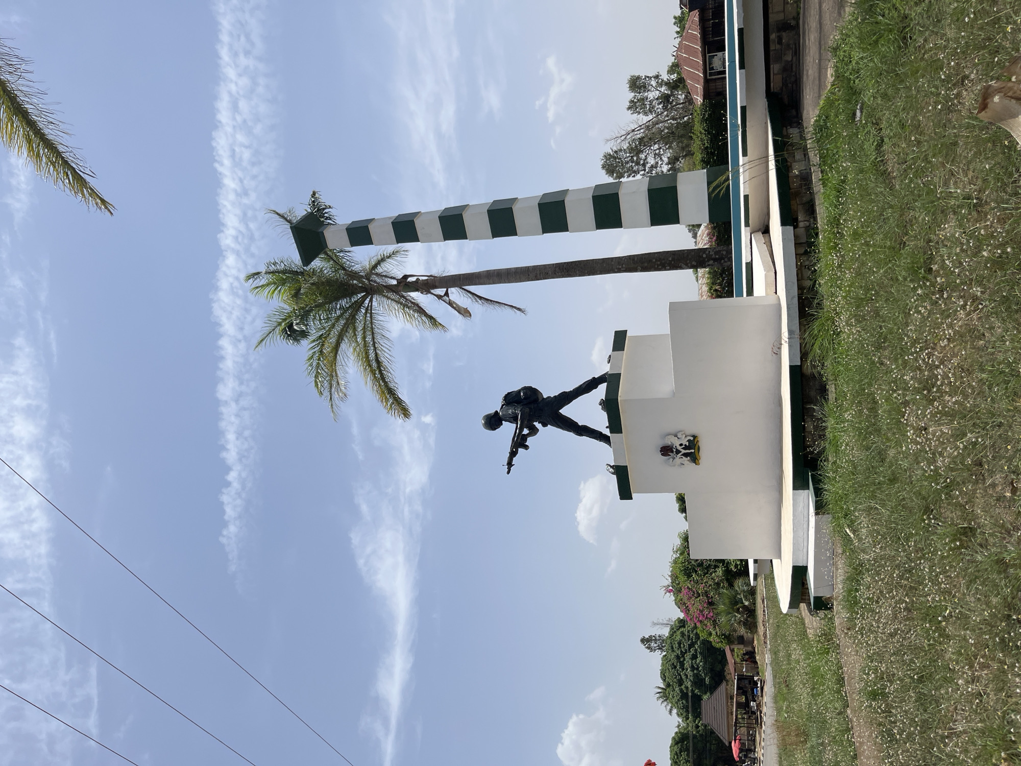 The image shows a war memorial in Jos, Nigeria. The statue depicts a soldier in a combat pose atop a white tiered base. Adjacent to the statue is a tall, cylindrical column painted with alternating green and white horizontal stripes. A palm tree is situated behind the memorial. The background includes a blue sky with scattered clouds and hints of surrounding vegetation. The image seems to be captured at ground level, presenting an upward perspective towards the memorial against the sky. A patch of grassy terrain is visible in the foreground.