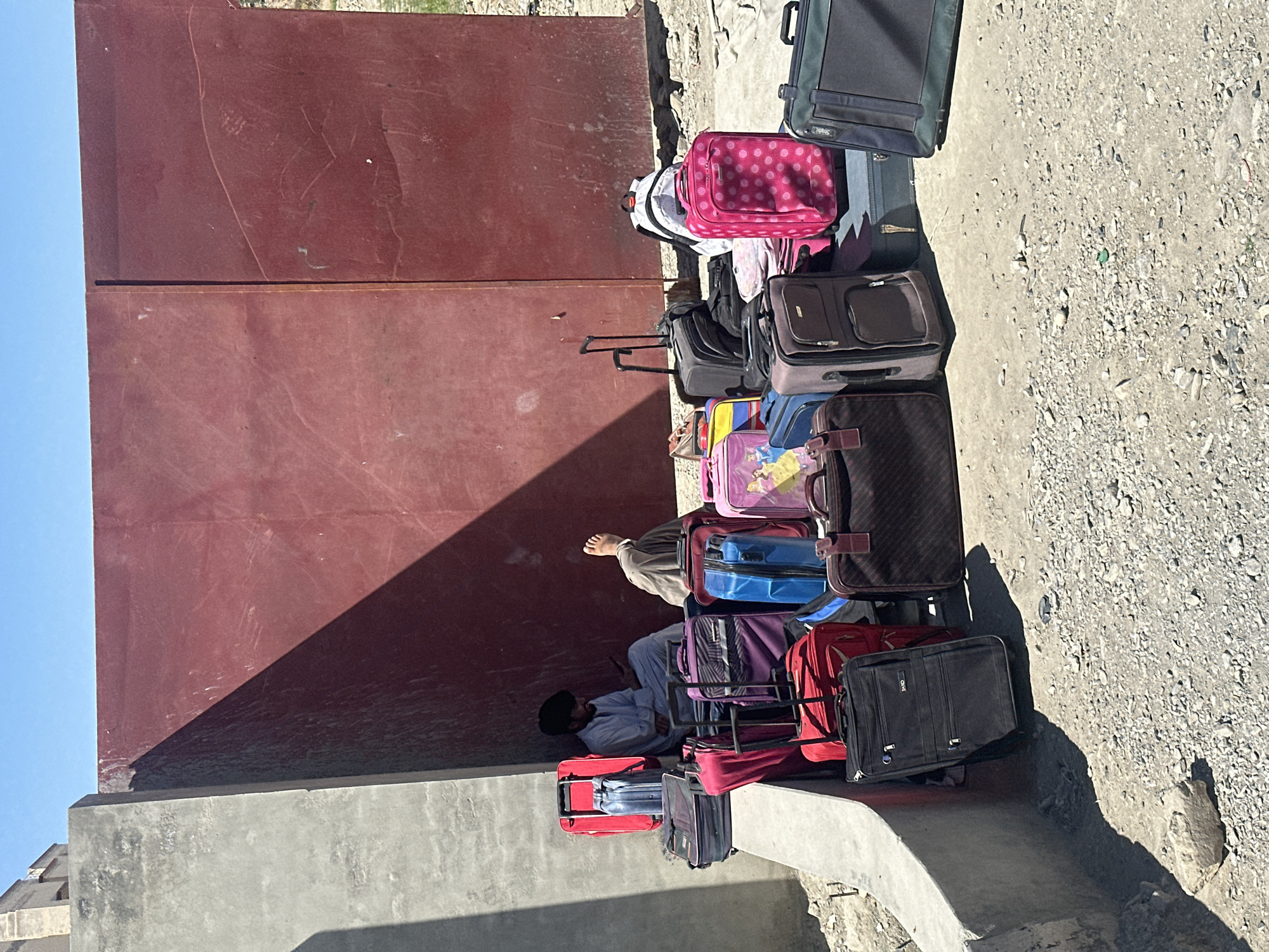 A row of luggage sits against a wall, mostly aligned in a relatively straight line. The luggage consists of various sizes, colors, and materials, including hard-sided suitcases and soft-sided bags. Among the luggage, a person is seen, wearing a light-colored outfit. The wall is a reddish-brown, likely made of metal or a similarly hard material. The adjacent ground is a light-colored, possibly concrete or stone surface, littered with debris. The environment appears arid and urban, possibly in a state of disrepair or construction. Based on the location, Quetta City Tehsil, Pakistan, it suggests an urban area with possibly infrastructure challenges.