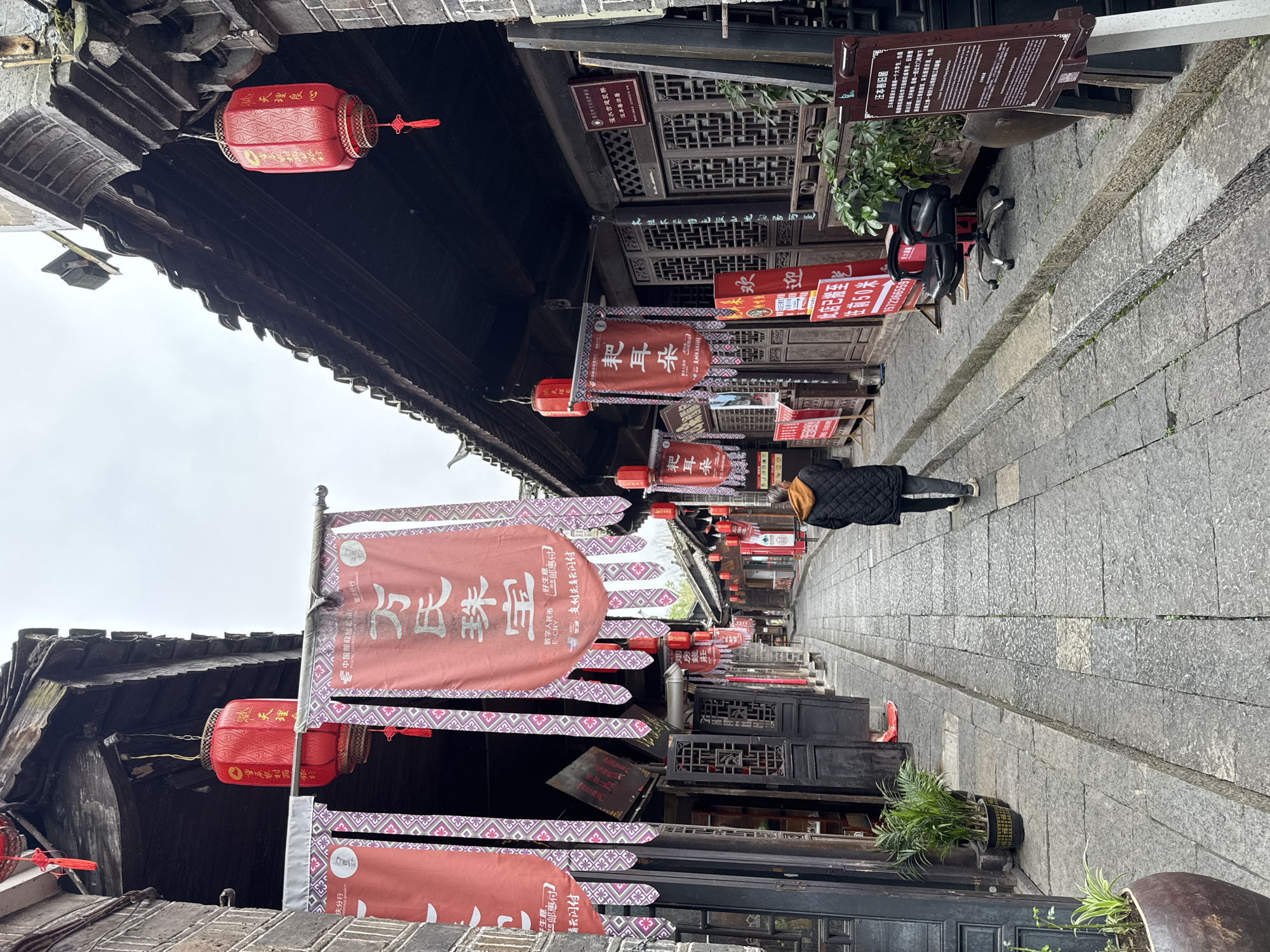 This is a street scene in Zhuoshui, China, featuring a narrow cobblestone pathway lined with traditional Chinese buildings. Red lanterns and banners with Chinese characters are strung overhead. A person wearing a black jacket walks down the street. A sign provides information about Wan's Jewelry. Another sign describes Wan Benshan's old residence. Office chairs sit by some plants.