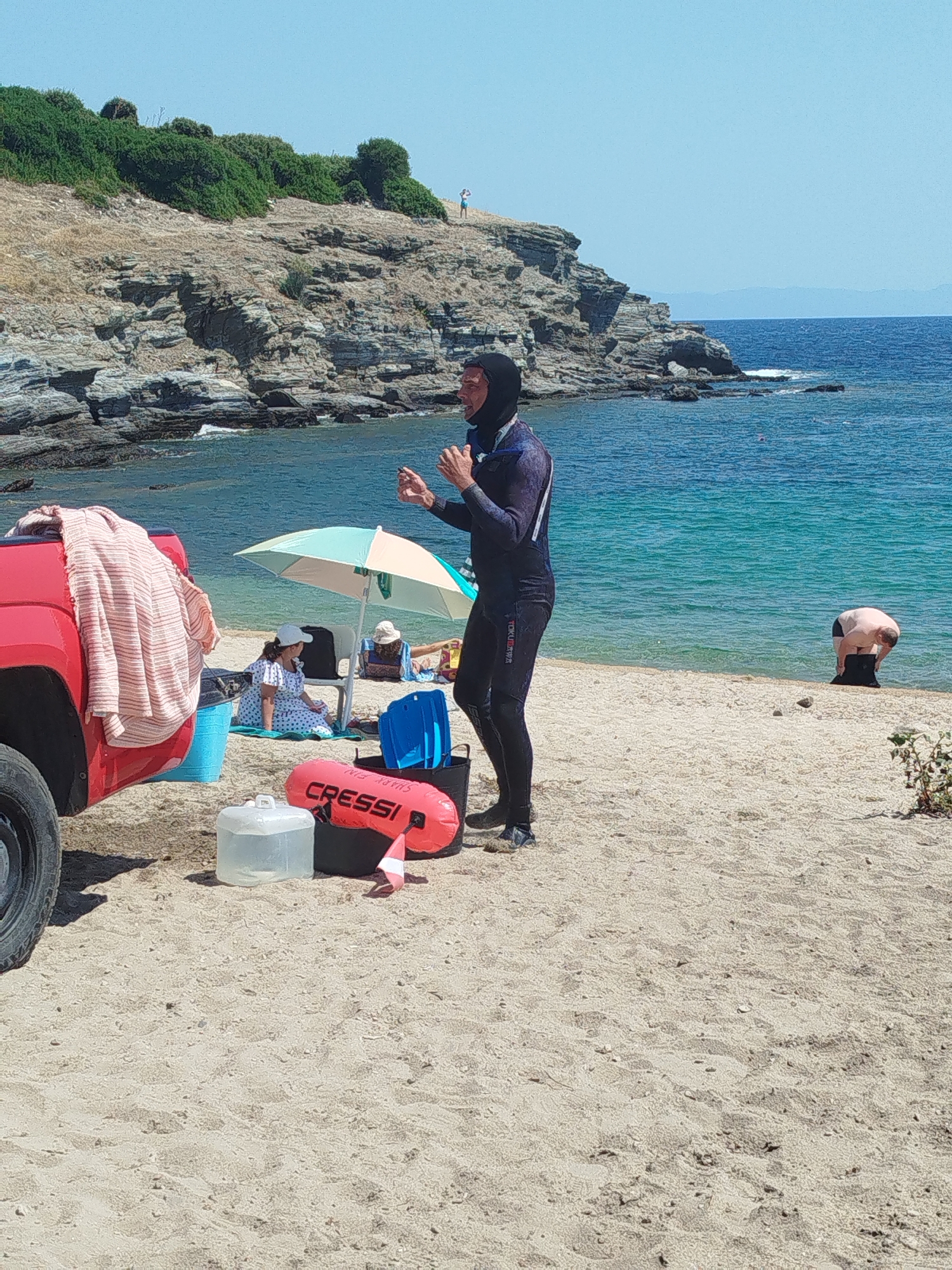 A group of people are on a sandy beach in the Sithonia Municipal Unit, Greece, near rocky cliffs and the blue sea. The scene includes a red pickup truck on the left with a pink and white striped towel draped over its side. In the center, a person in a black wetsuit is standing and gesturing. Nearby, there are beach chairs, a beach umbrella, and various beach items, including a red float with the word "CRESSI" on it, a blue bucket, and a clear plastic water jug. Two people are sitting under the umbrella, one wearing a white hat and a polka-dot dress, and another person is partially visible lying down. Further down the beach, a man is bending over near the water's edge, appearing to be putting on or taking off a black bag. The sky is clear and blue, indicating a sunny day. The environment is a coastal area with vegetation on the rocky outcrops.