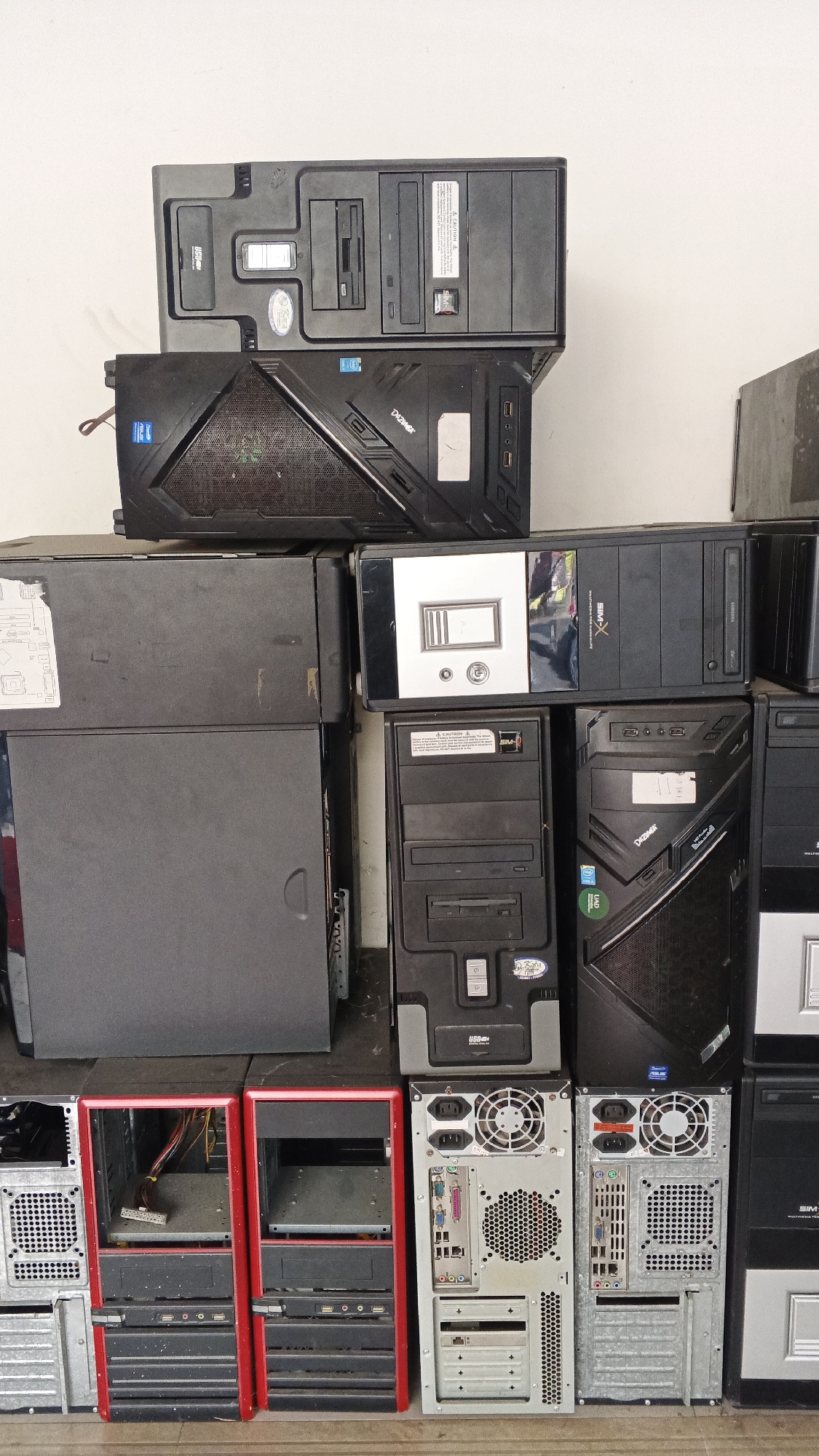 The image shows a stack of computer towers in what appears to be a storage area or a repair shop. The towers are of various shapes, sizes, and colors, with some being black, gray, and red. Some of the towers are open, revealing internal components like wires and fans. The environment appears to be indoors, with a white wall in the background. There are no people or animals visible in the image. The scene is cluttered, with the computer towers occupying most of the frame. The location is identified as Banguntapan, Indonesia, suggesting this is a scene from that region.