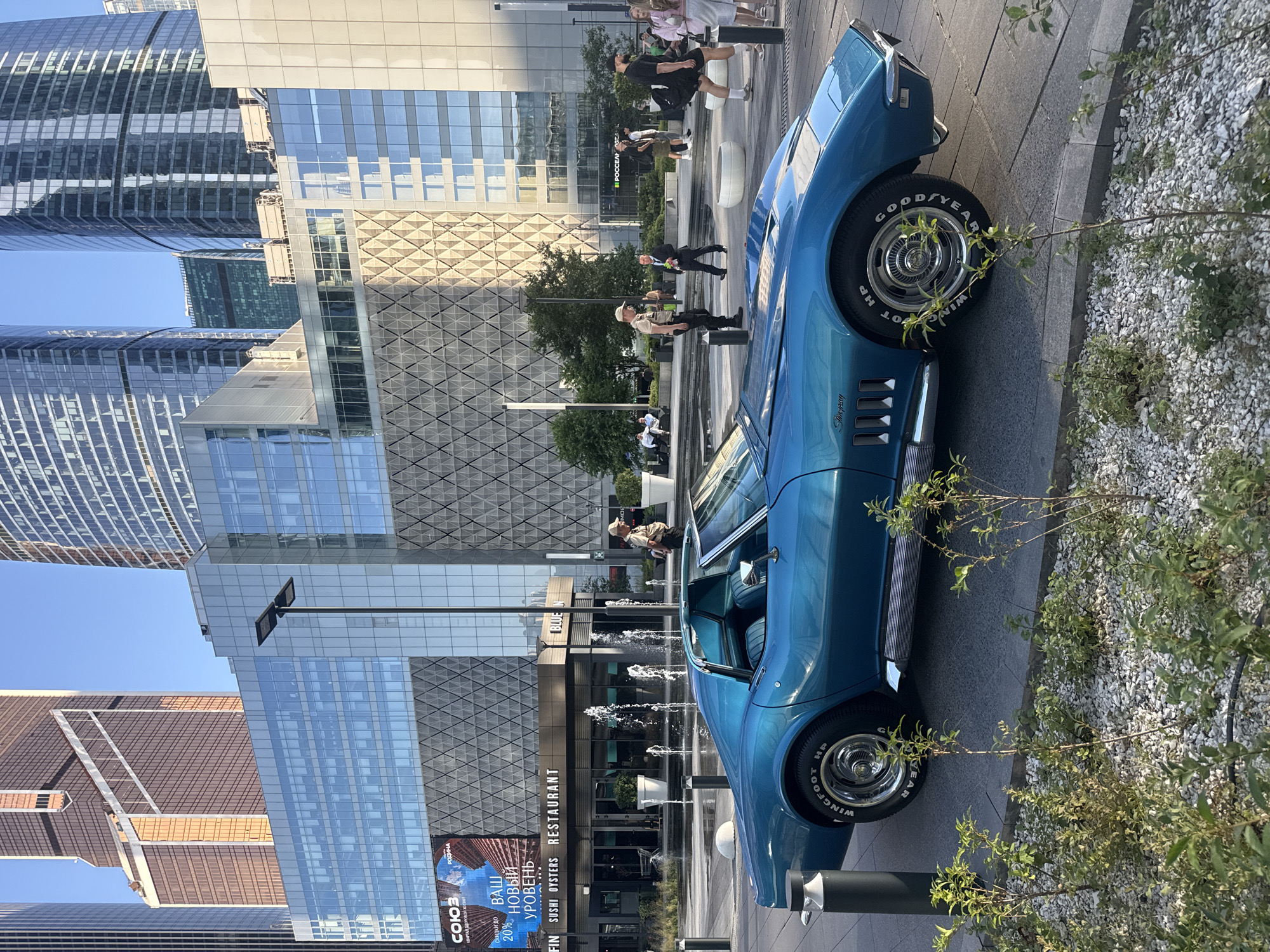 The image depicts a blue Chevrolet Corvette Stingray parked in front of a modern building complex. The architecture suggests a contemporary urban environment, characterized by glass facades and towering skyscrapers. In the background, a large digital billboard displays an advertisement, and a fountain with water jets is visible. Several people are present in the scene, some walking, others seated. The car's classic design contrasts with the modern surroundings. The foreground features a patch of low-lying greenery and gravel. The overall impression is that of a stylish vehicle showcased in a bustling, upscale urban setting.