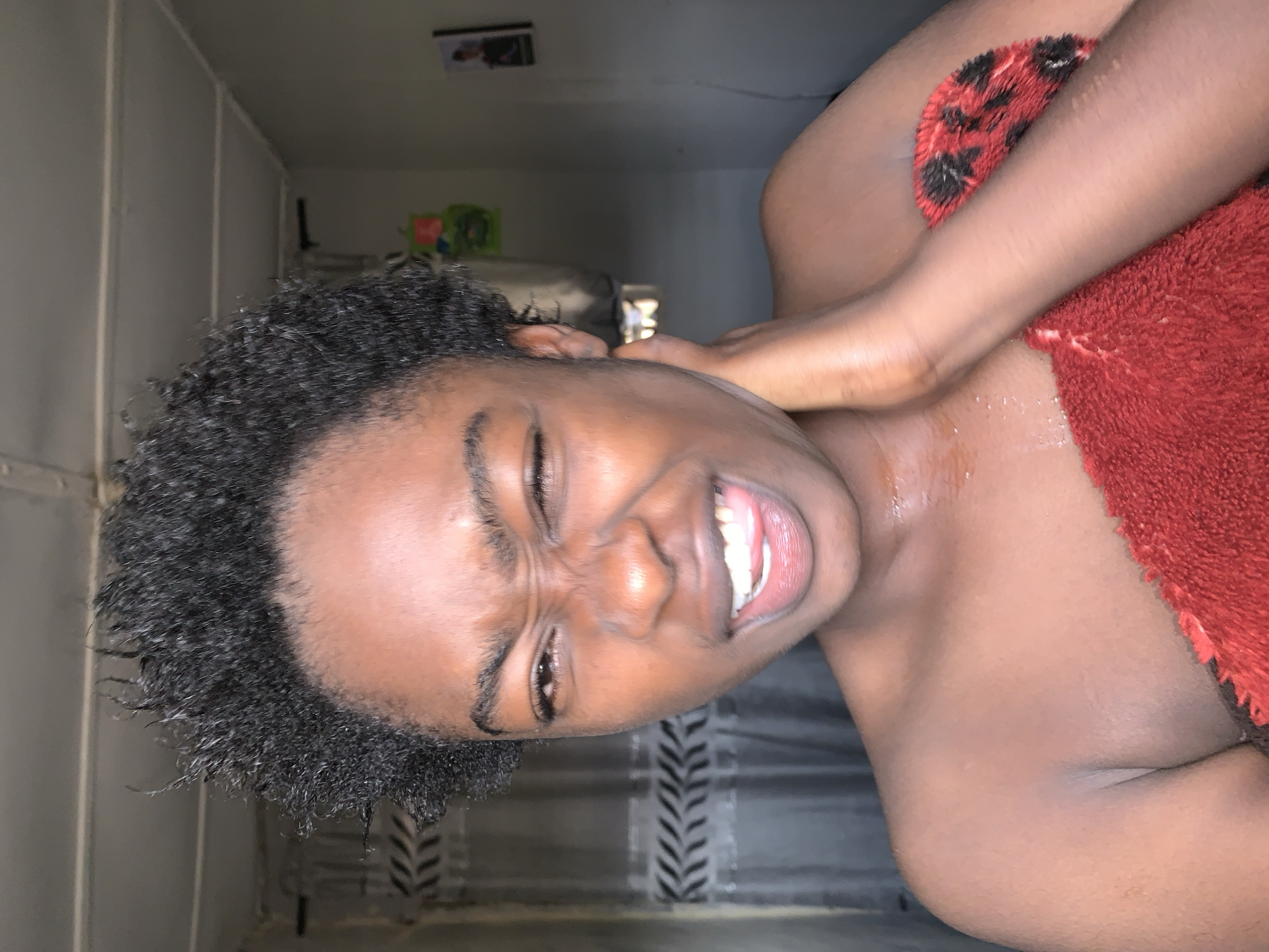 A person with dark curly hair is smiling at the camera with one eye closed. A red towel is draped over their shoulder. The background appears to be an interior space with gray walls. The image is vertically oriented. There are no visible elements to identify the location as Rafin Par, Nigeria.
