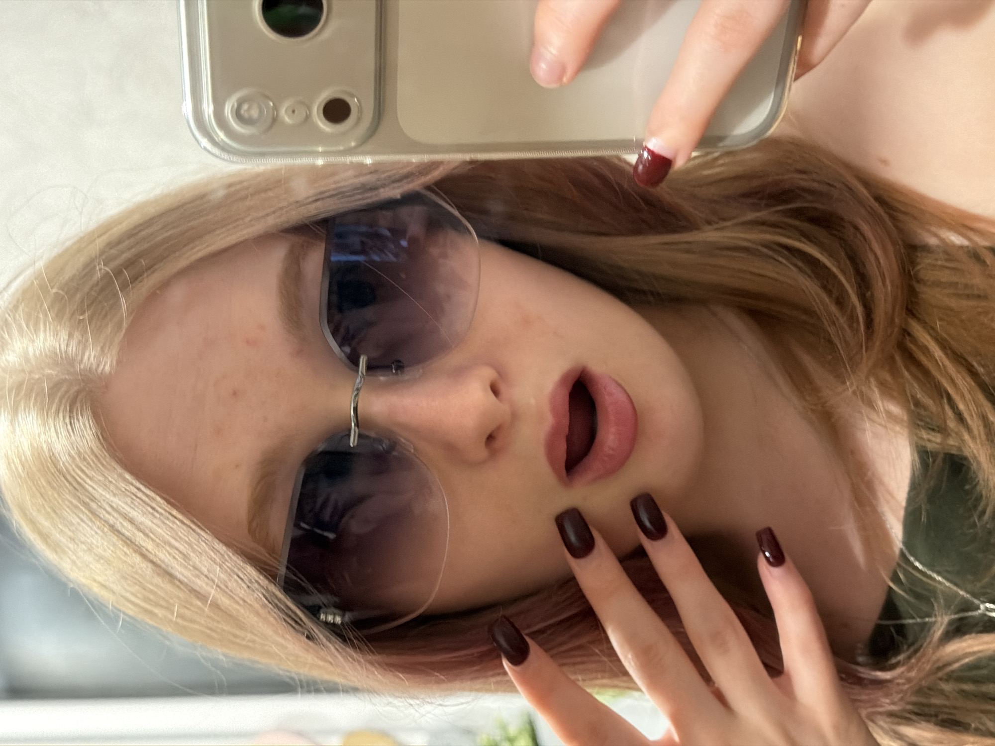 A fair-skinned woman with blonde and auburn hair is taking a selfie with a phone in a clear case. She is wearing rimless sunglasses. Her nails are painted a dark maroon color. The location is unknown and cannot be determined from the image.