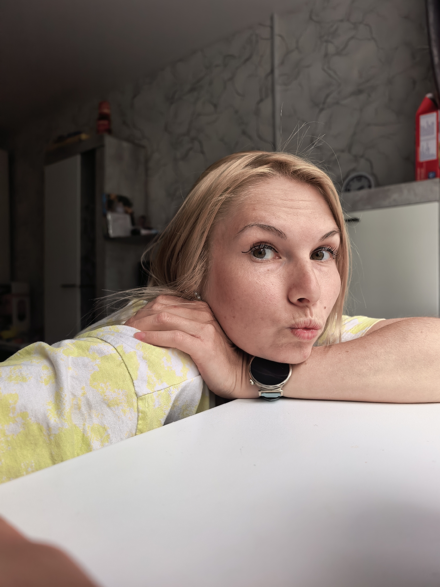 A fair-skinned woman with blonde hair is posing for a selfie. She is resting her cheek on her hands, which are on a white surface, possibly a table. She has a smartwatch on her left wrist. She is making a kissing face at the camera. The background shows a kitchen interior with a cabinet and patterned wallpaper. A red carton is visible on the shelf. No specific location in Sterlitamak, Russia, is discernible from the image.