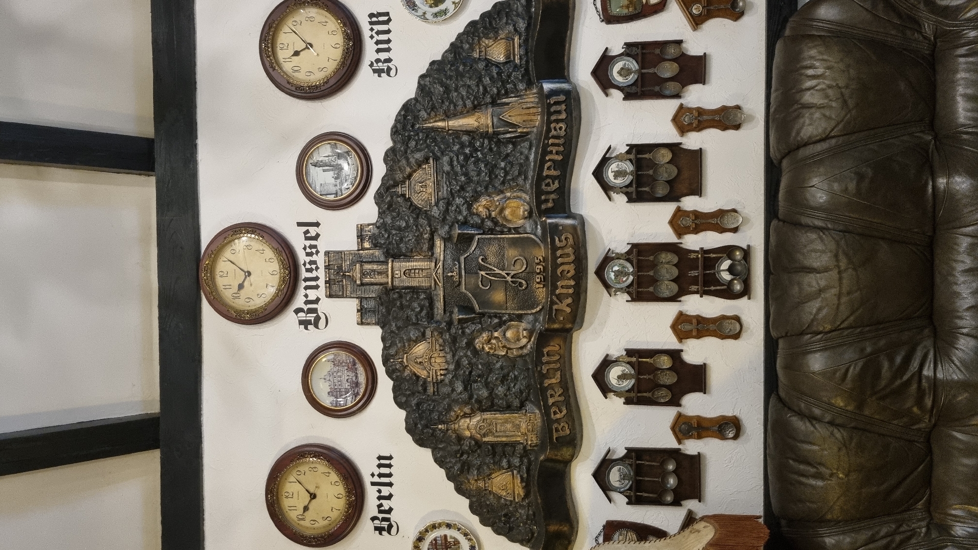 The image shows a wall decorated with several clocks and a large, ornate bas-relief featuring a castle and the words "Berlin Knaus Чернівці" along with the year "1995". The bas-relief also includes architectural elements and shields. Below the bas-relief are several wooden wall decorations, each holding numerous decorative spoons. The wall appears to be white and textured, with a dark wooden beam running across the top. In front of the wall, a large, dark brown leather sofa is visible. The scene is indoors, likely a room or establishment in Chernivtsi, Ukraine, given the inscription. There are no people visible in the image.
