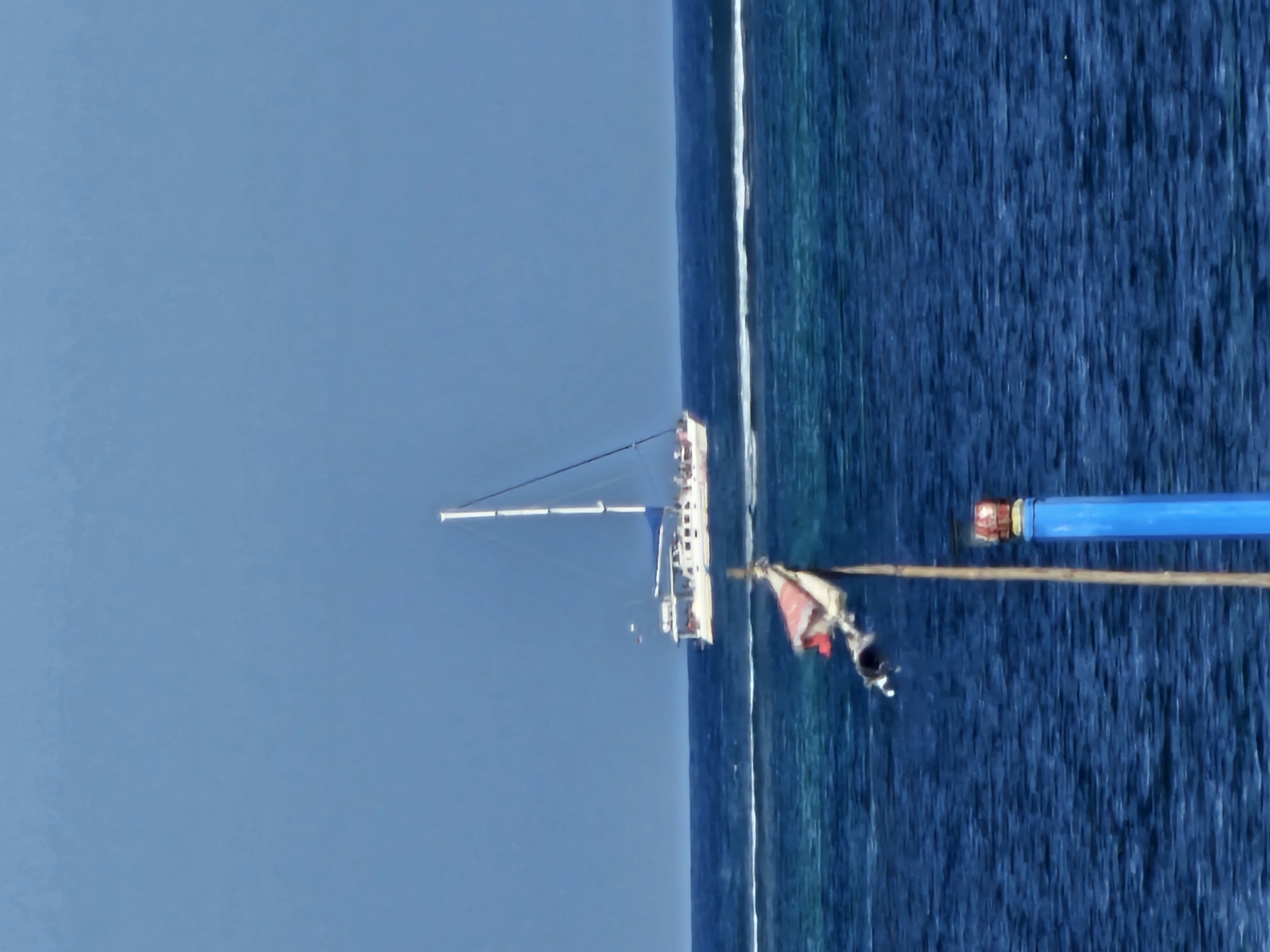 The image depicts a serene ocean scene, likely near Denpasar, Indonesia, given the context. The vast expanse of the deep blue sea dominates the frame, meeting the lighter blue sky in a clear horizon. A white sailboat with a tall mast is a prominent object, positioned in the mid-ground, appearing to be at anchor or moving slowly. Its sails are partially furled, suggesting a calm day.
In the foreground, a rough-looking wooden pole extends out over the water, adorned with a tattered red and white flag that flutters slightly. Attached to the pole's end is a bright blue cylindrical object, possibly a bu oy or part of some equipment, with a red cap visible on its end. The water appears clear, with hints of lighter turquoise near the shore, indicating shallow coral reefs or sandbanks.
The overall impression is one of a peaceful day at sea, possibly mid-morning or late afternoon given the soft lighting and clear skies. There are no discernible people clearly visible on the sailboat or in the water, though the flag's movement might suggest a light breeze. The scene conveys a sense of tranquility and isolation, typical of a tropical coastal environment. No text is visible in the image.
