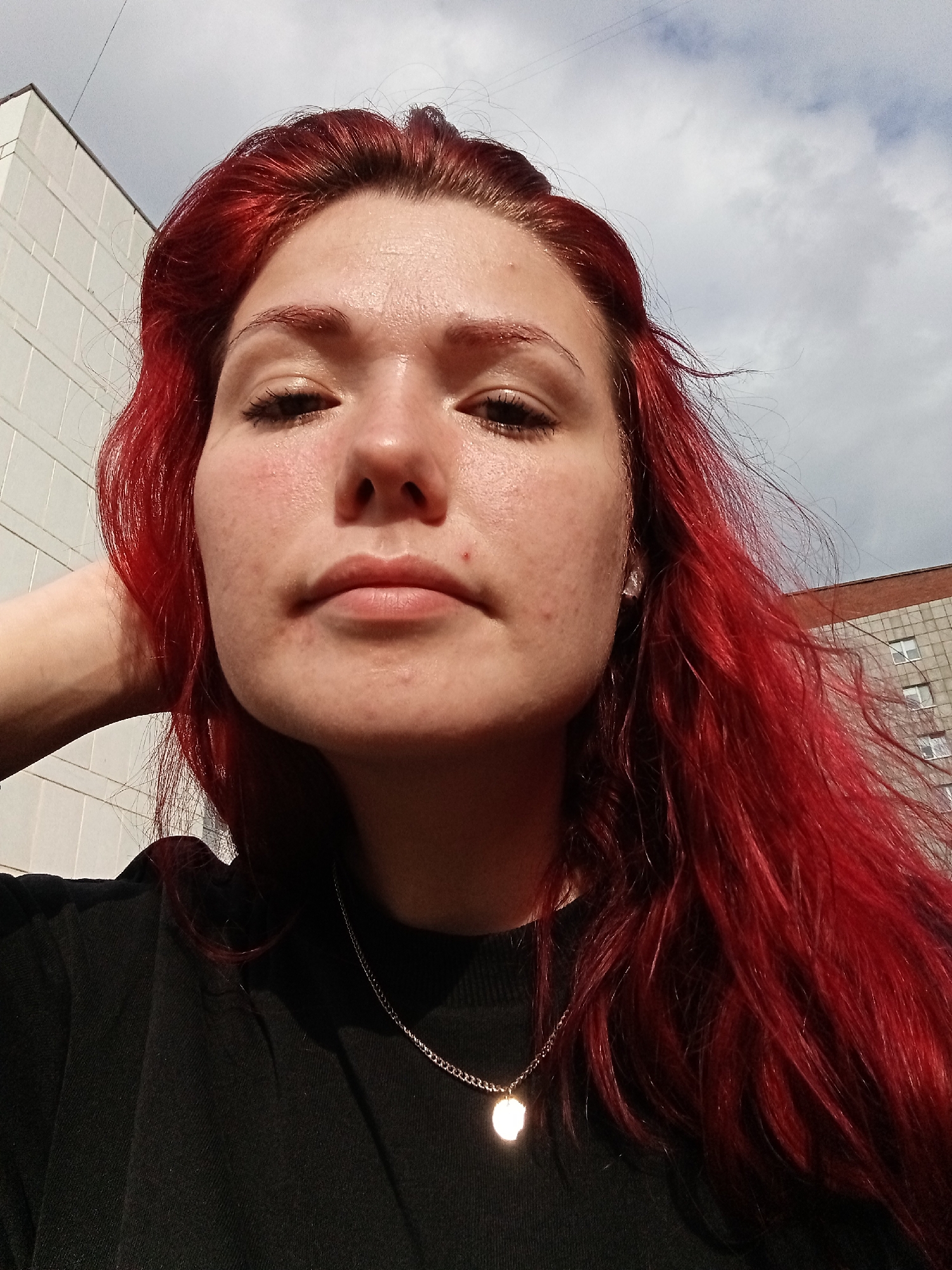 A young woman with bright red hair is taking a close-up selfie outdoors. She is wearing a black t-shirt and a silver chain necklace with a round pendant. Her left arm is bent, with her hand resting behind her head. Her skin appears slightly flushed, and she has some minor blemishes on her face. The background shows a cloudy sky, a white building with horizontal lines, and a brick building with windows. The overall lighting suggests it is daytime. This image appears to have been taken in Chaikovsky, Russia, possibly near residential buildings characteristic of the city.