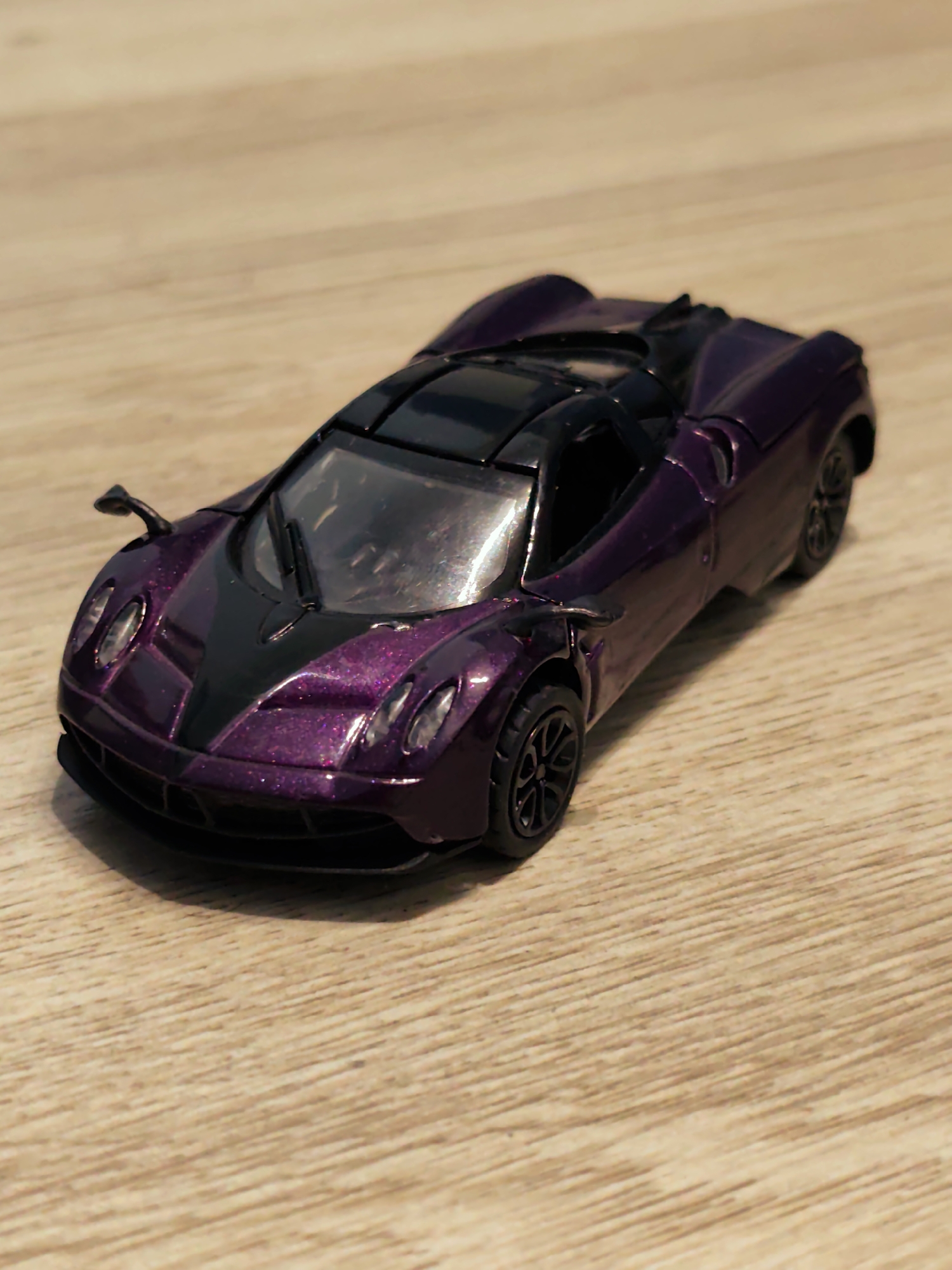 A die-cast model of a high-performance sports car is positioned diagonally on a light-colored wooden surface. The car's exterior is a metallic purple with visible glitter particles, contrasting with matte black sections on the central front hood, roof, and lower side sills. The model features detailed elements including headlights, side mirrors, and a single central windshield wiper. Its wheels are black with a multi-spoke design. The wooden surface, serving as the background, displays a horizontal grain pattern. The lighting source creates reflections on the car's glossy finish. No other objects or living beings are visible in the scene. The image contains no discernible visual cues or identifying marks to indicate a specific geographical location, such as the city of Pazardzhik, Bulgaria.