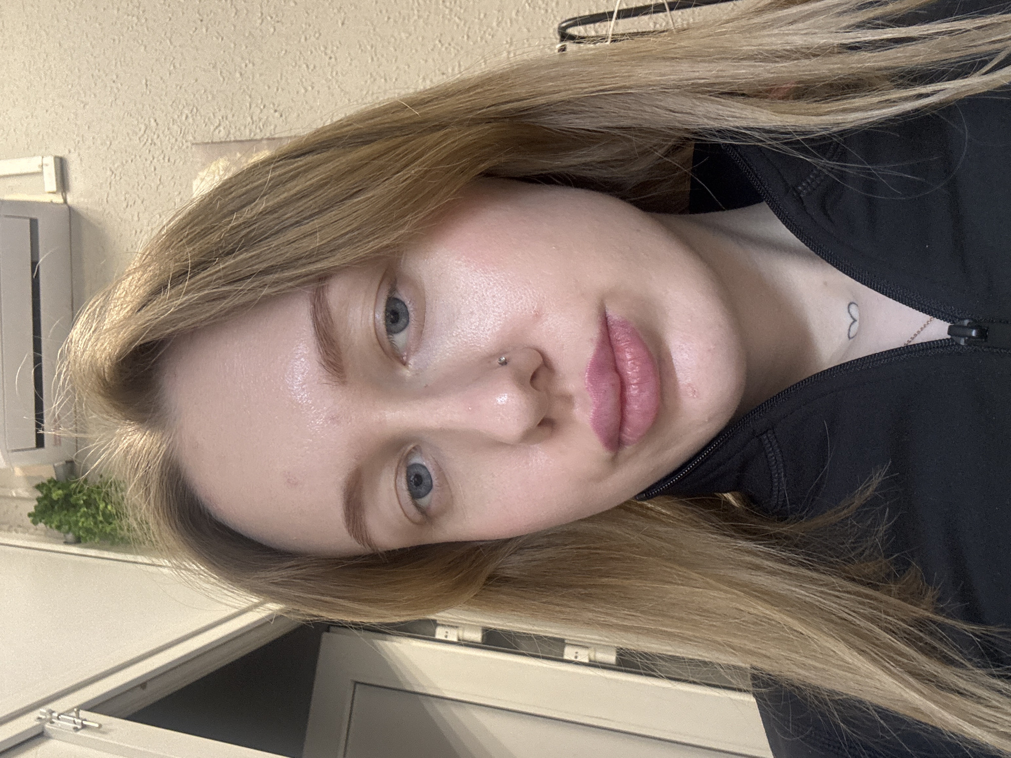 A close-up shot of a woman with blonde hair and blue eyes. She is wearing a black jacket with a partially visible zipper. A small heart tattoo is visible on her chest. She has a nose piercing. The location is difficult to discern from the image; there is no visible indication that it is Rostov-on-Don, Russia. The background is a light beige wall and a white door frame. A small plant is visible on a surface behind the woman.
