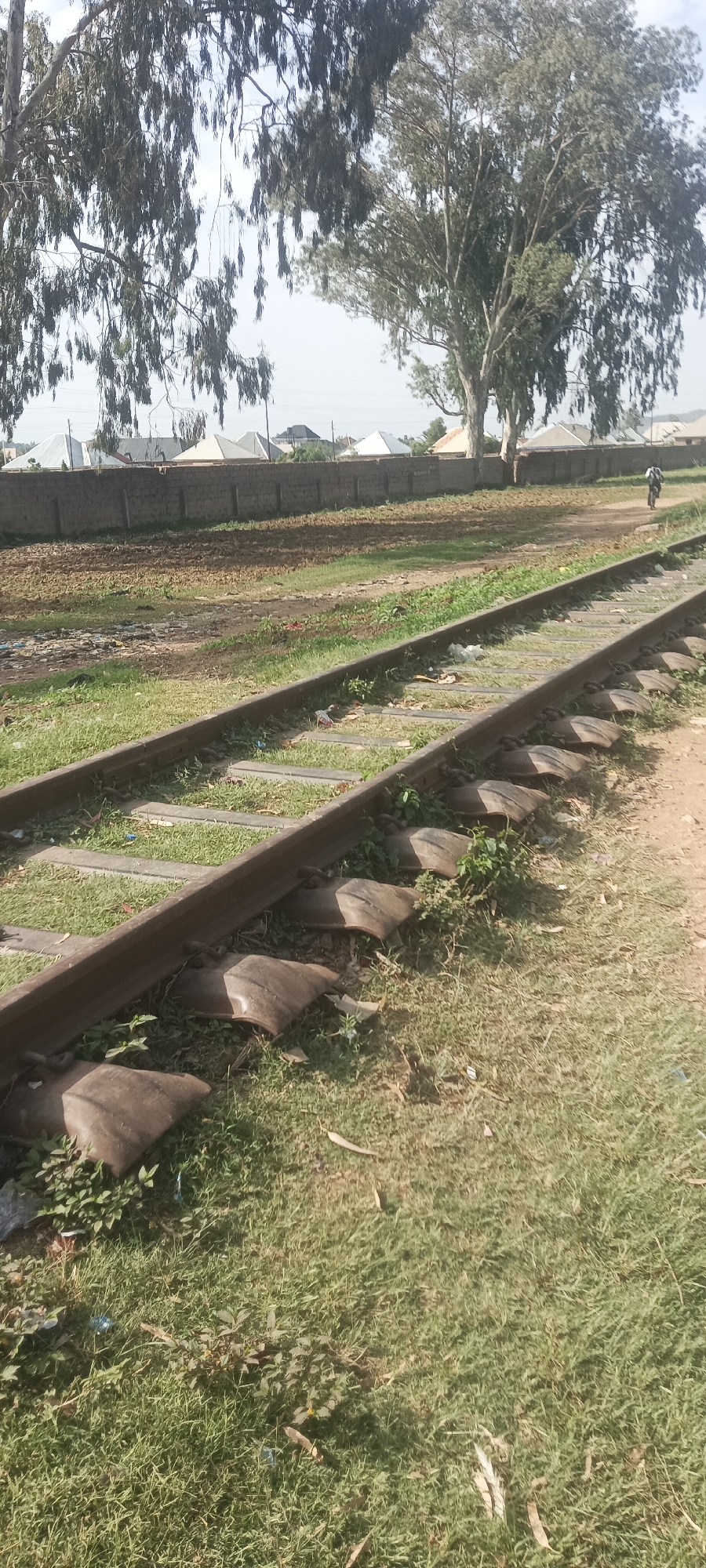 The image depicts a section of railway tracks in an apparent open area within the city of Bukuru, Nigeria. The tracks, showing signs of disuse, are set into the ground with rudimentary, worn-out tire-based sleepers instead of traditional railroad ties. Vegetation is growing around and through the tracks. In the background, there is a low concrete or stone wall with residential buildings of a similar design beyond the wall, indicating an urban setting. One figure is visible in the far background, appearing to be on a bicycle or motorbike. The overall environment suggests neglect or abandonment of the rail infrastructure.