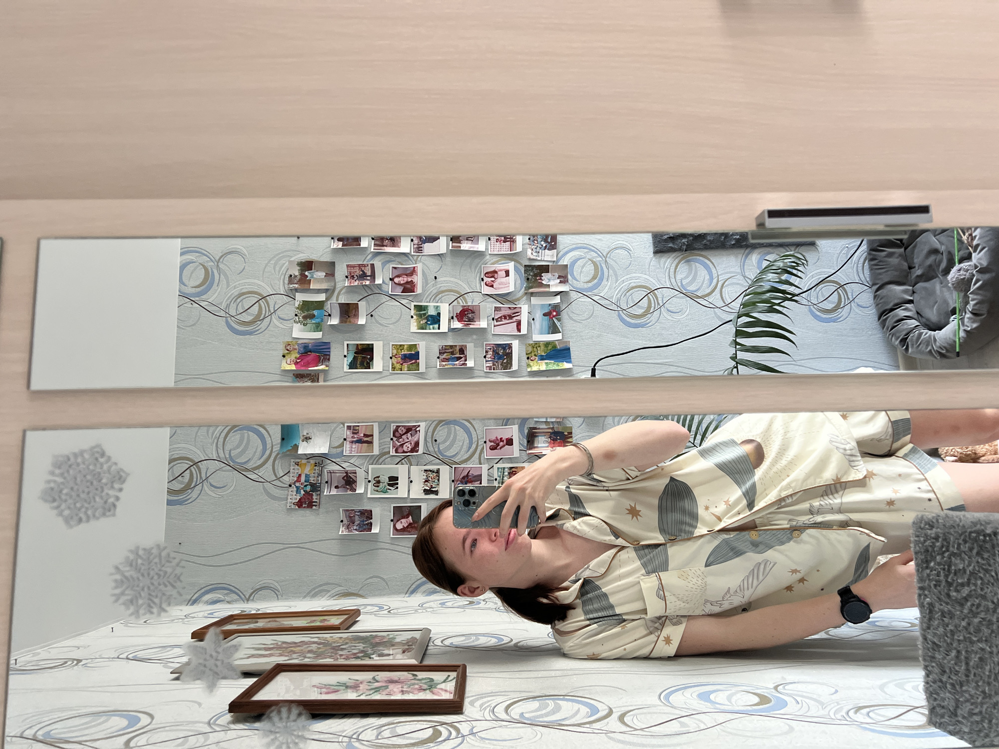 A woman is lying on a bed, taking a selfie in a mirror. She is wearing a patterned pajama set and a watch. The bed has a patterned duvet, and there are framed pictures on it. The wall behind the bed is decorated with wallpaper featuring a swirling pattern and photographs attached with string. A plant and a grey cushion are also visible. The location is Chaikovsky, Russia.