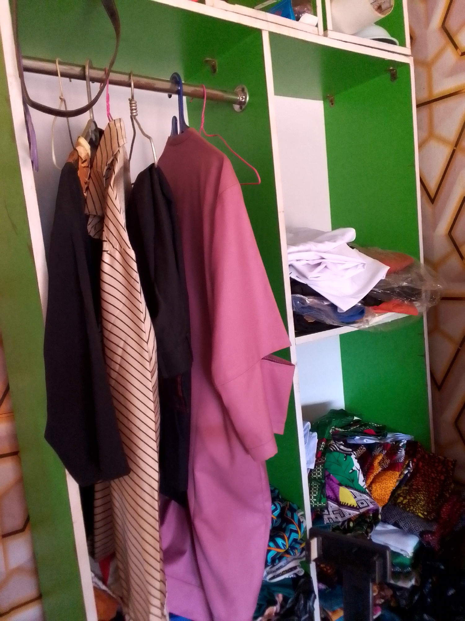 A medium shot shows a green and white wardrobe with clothes hanging in the foreground, possibly in Unknown, Unknown.
Hanging on a horizontal rod are three garments of different colors and patterns. On the left is a black garment, followed by a tan and black striped garment, and a mauve garment on the right.
Inside the wardrobe, stacked neatly, are a pile of folded white shirts, some in plastic packaging, on the middle shelf. The bottom shelf reveals a disorganized assortment of colorful fabrics.
The wardrobe has green sides and a white interior. A wall with a geometric pattern is visible behind the wardrobe.