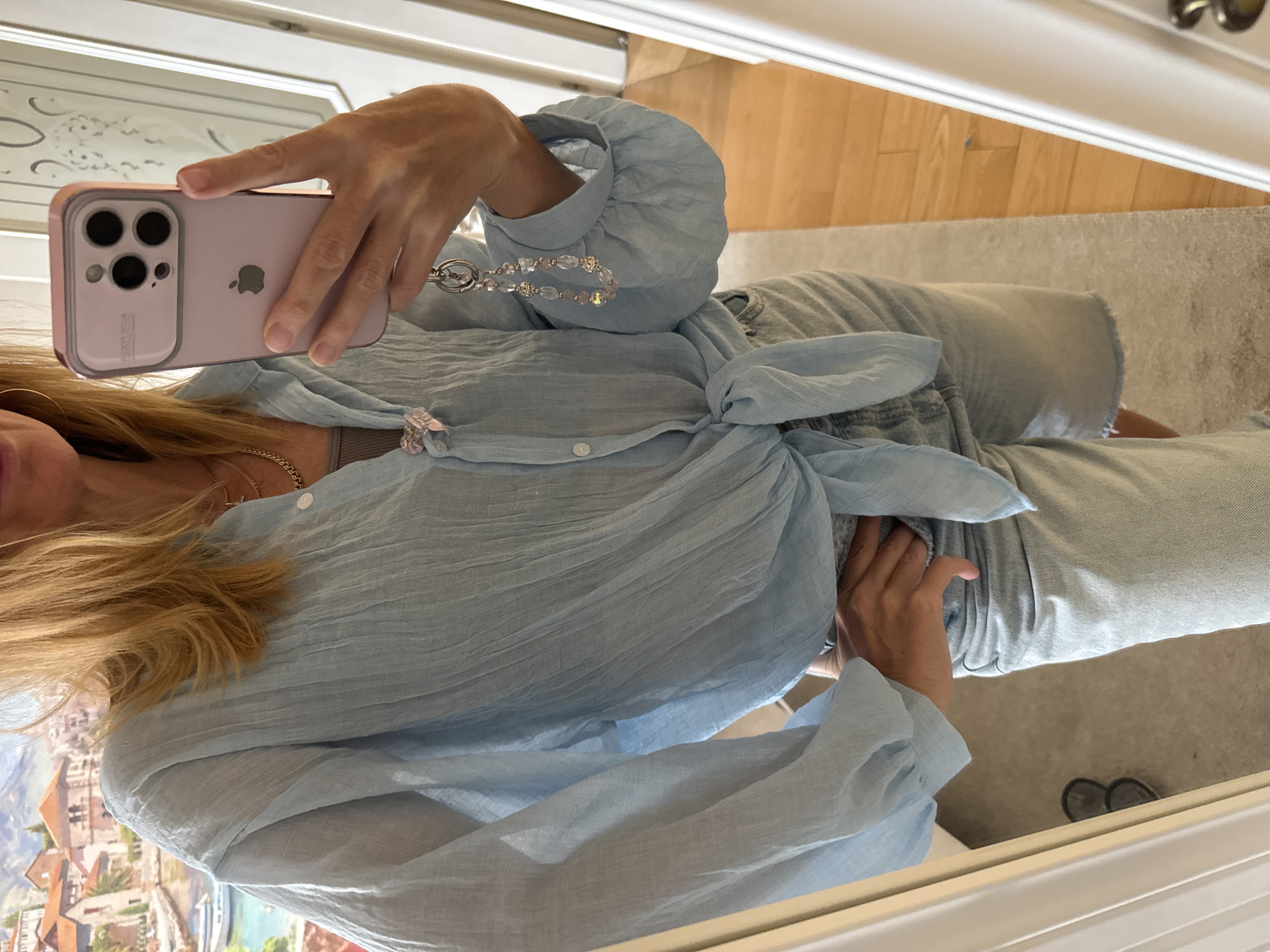 A person is taking a mirror selfie, wearing a light blue, crinkled long-sleeved shirt tied at the waist and light wash denim shorts. They are holding a light purple phone in their right hand, which has a keychain attached. A pink bow adorns the front of the shirt. The background shows a wall with a painting of a Mediterranean-style village with a body of water and boats, and part of a doorway. The floor appears to be wood flooring. The location is identified as Unknown, Russia.